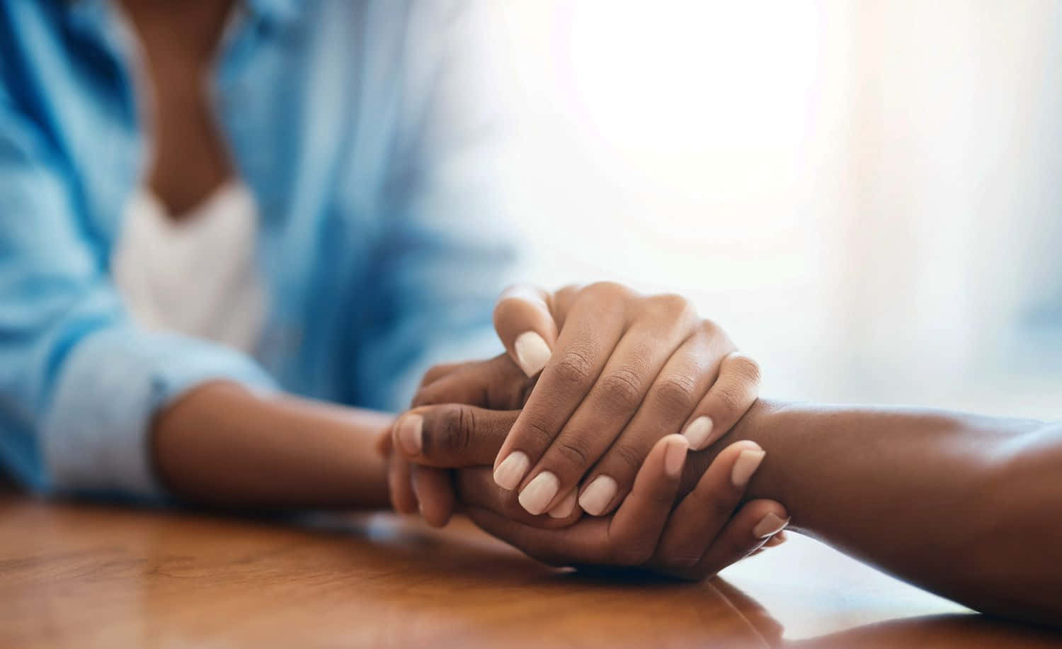 Holding Hands 桌布 壁紙 — 兩隻手帶著淺粉紅色指甲，輕輕交握在一張木桌上，處於柔和照明的室內環境中。