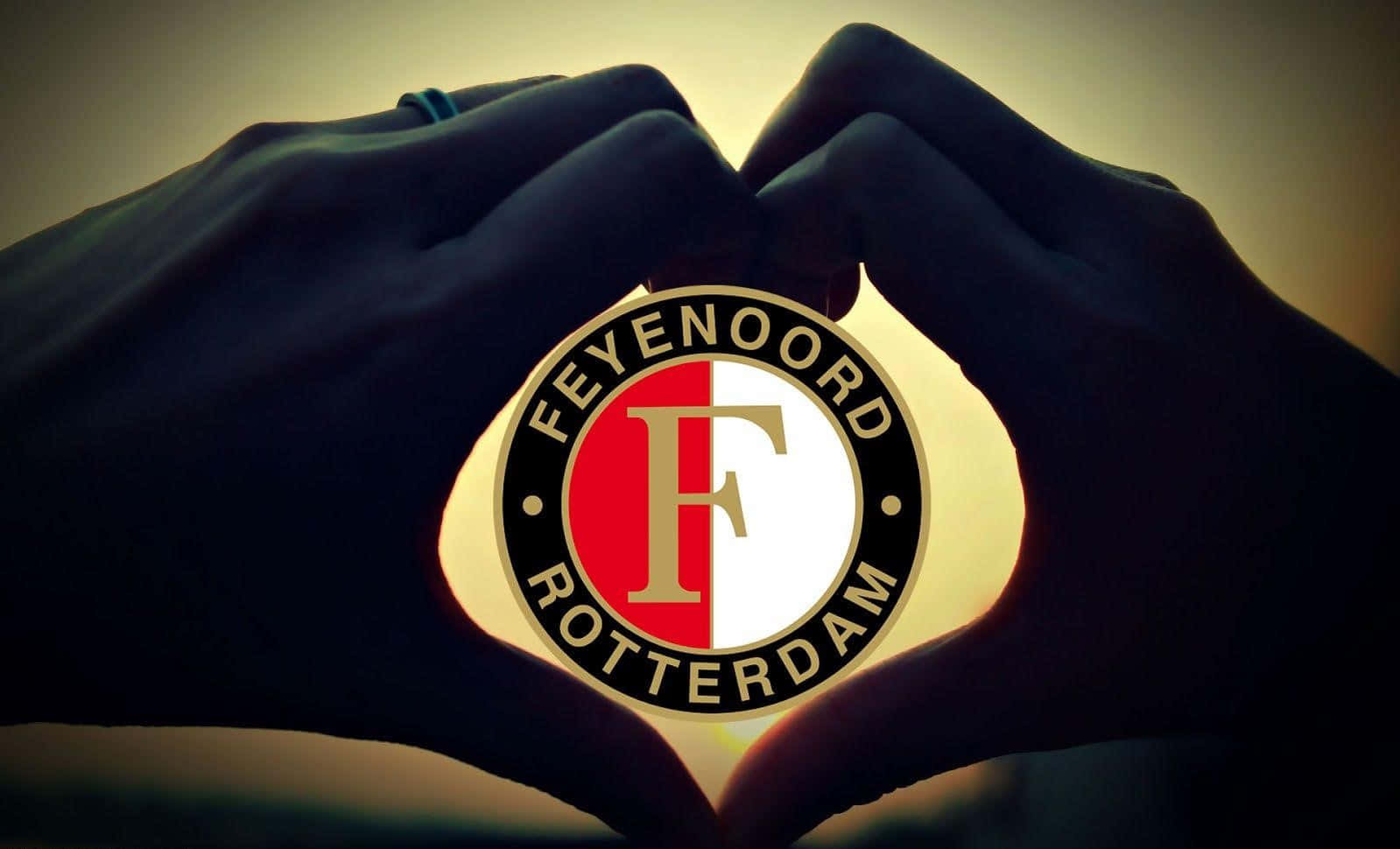 Feyenoord 桌布 壁紙 — 兩隻手形成心形，框住發光的飛燕諾鹿特丹足球俱樂部徽章，背景為溫暖的漸變色。