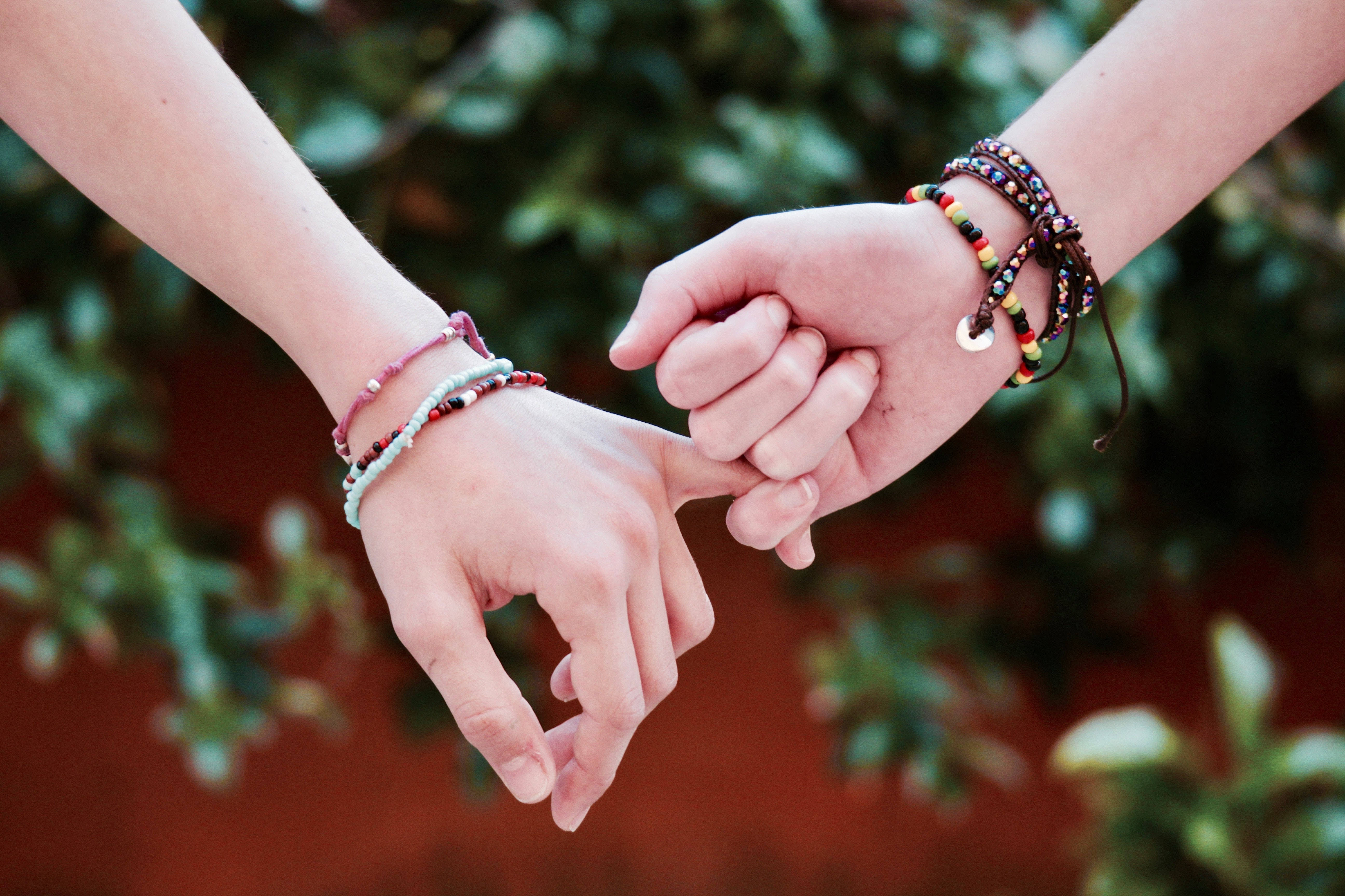 pinky promise 桌布 壁紙 — 兩隻手戴著色彩繽紛的珠串手鐲，在柔和模糊的綠色和陶土色背景前輕輕相握。
