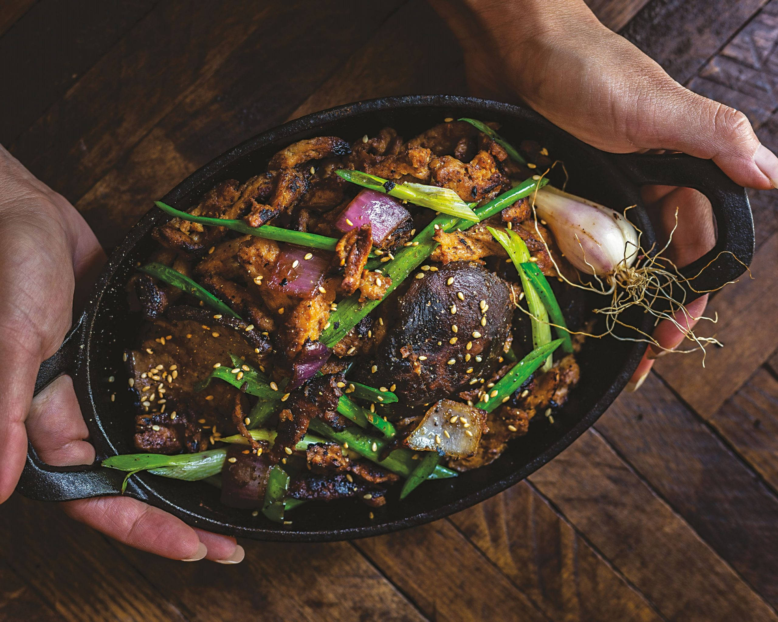 meat 桌布 壁紙 — 兩隻手拿著一個鑄鐵煎鍋，裡面裝滿了滋滋作響的辣味炒菜，上面撒有蔥花、芝麻和整個蒜頭，放在一個質樸的木桌上。