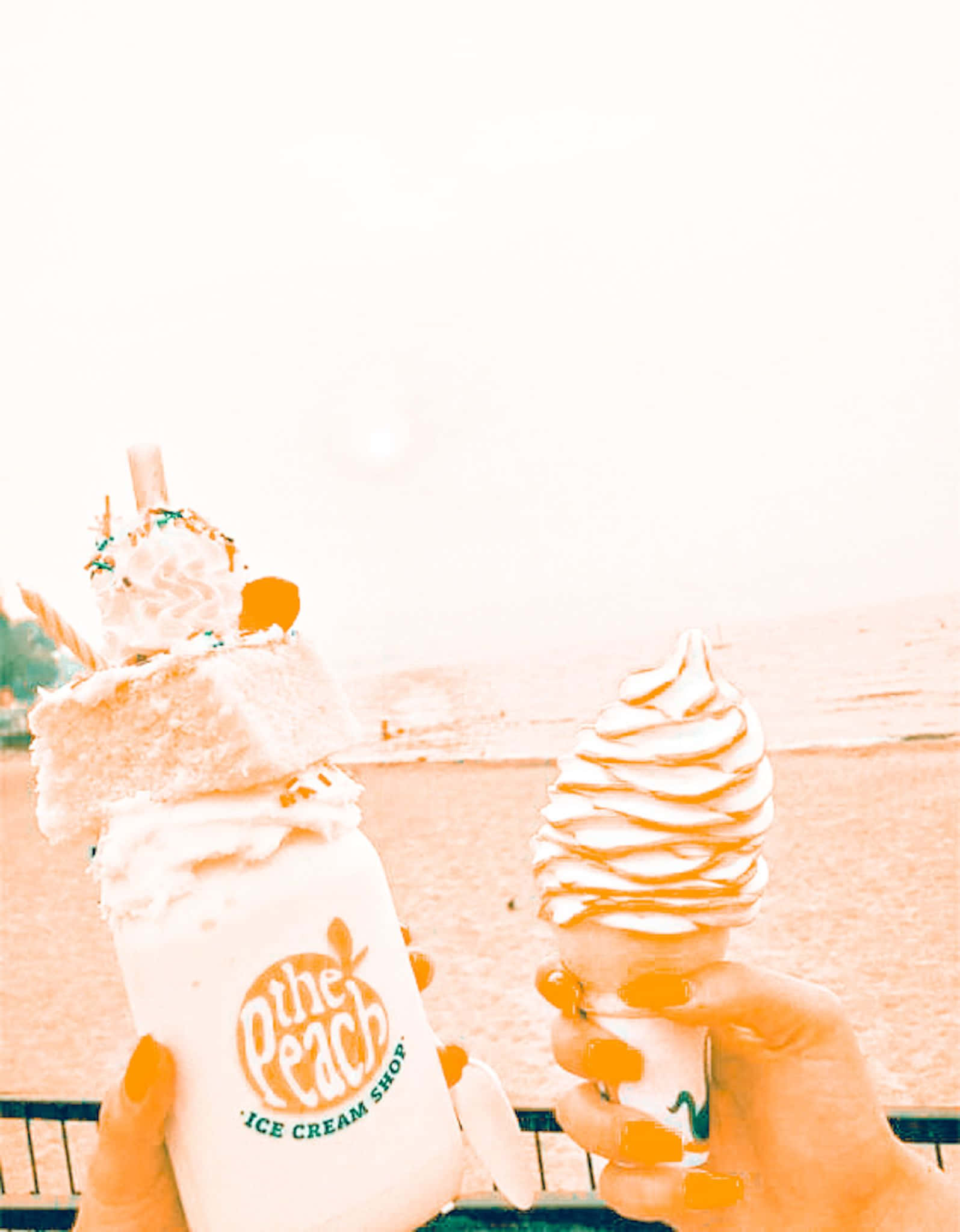 Peachy Aesthetic 桌布 壁紙 — 兩隻手拿著一個高聳的蜜桃冰淇淋聖代和一個漩渦甜筒，背景是朦朧的海灘，帶有溫暖的橙色調。