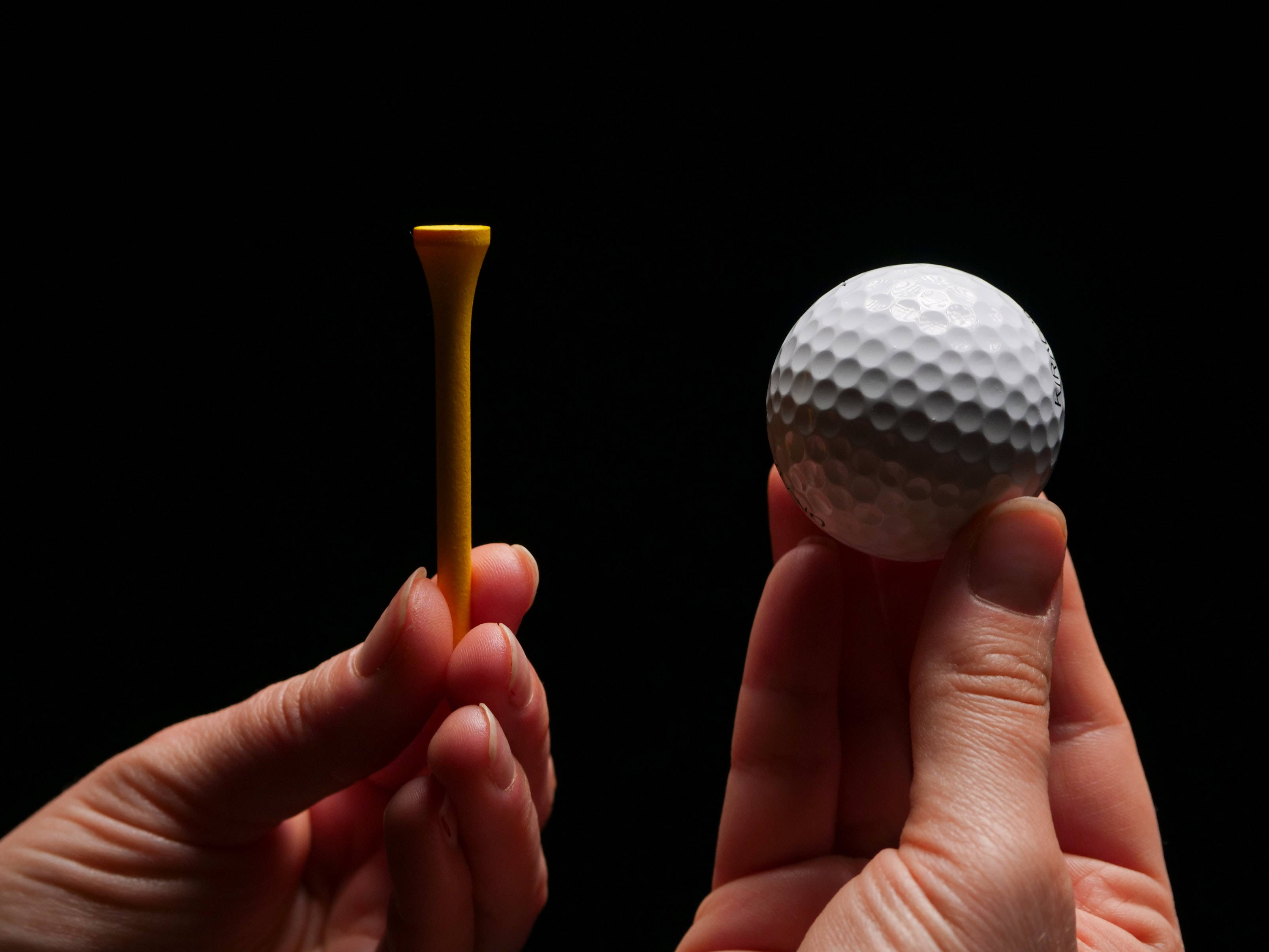 4K Golf 桌布 壁紙 — 兩隻手拿著一個黃色高爾夫球座和一個白色帶凹點的高爾夫球，背景是 stark 黑色。