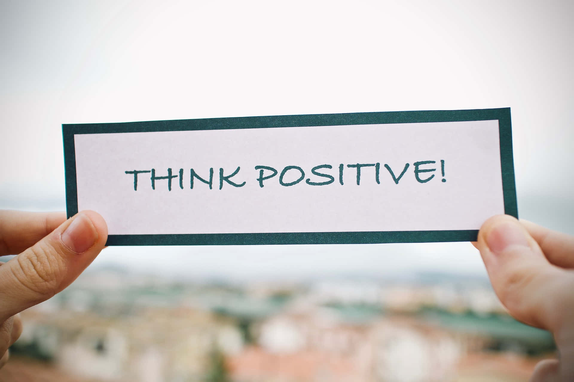 Reminder 桌布 壁紙 — 兩隻手拿著一張手寫便條，上面寫著「THINK POSITIVE!」，背景是模糊的城市景觀。