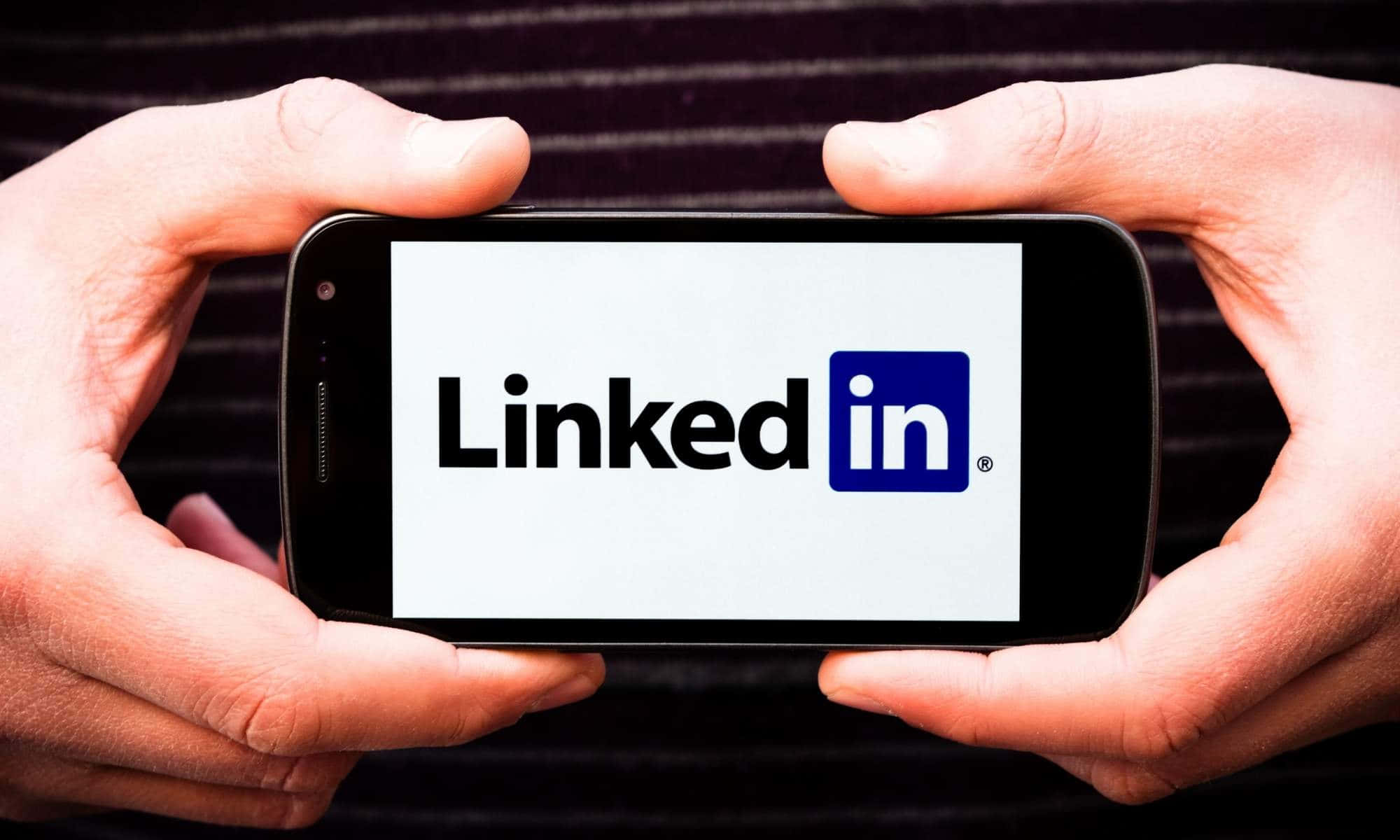 Best Linkedin 桌布 壁紙 — 兩隻手拿著一支智慧型手機，顯示LinkedIn標誌，背景為深色條紋。