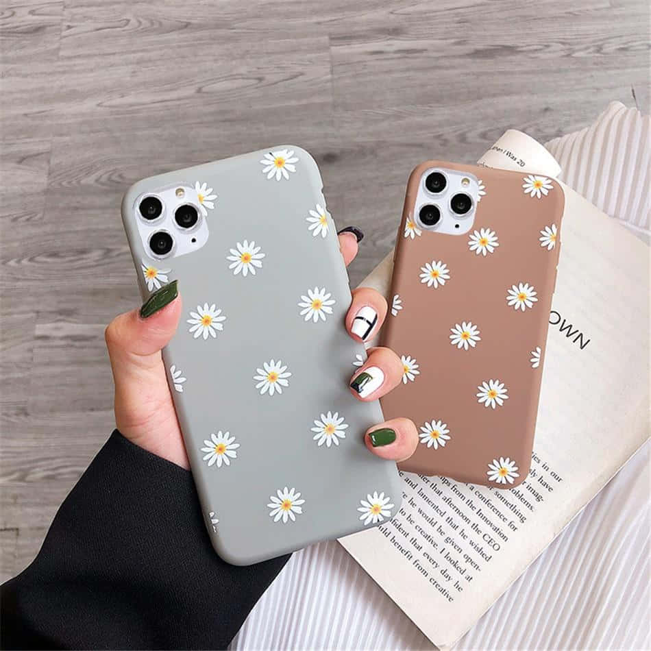 Phone Case 桌布 壁紙 — 兩隻手拿著帶有白色雛菊圖案的粉彩手機殼，背景是木地板和一本打開的書。