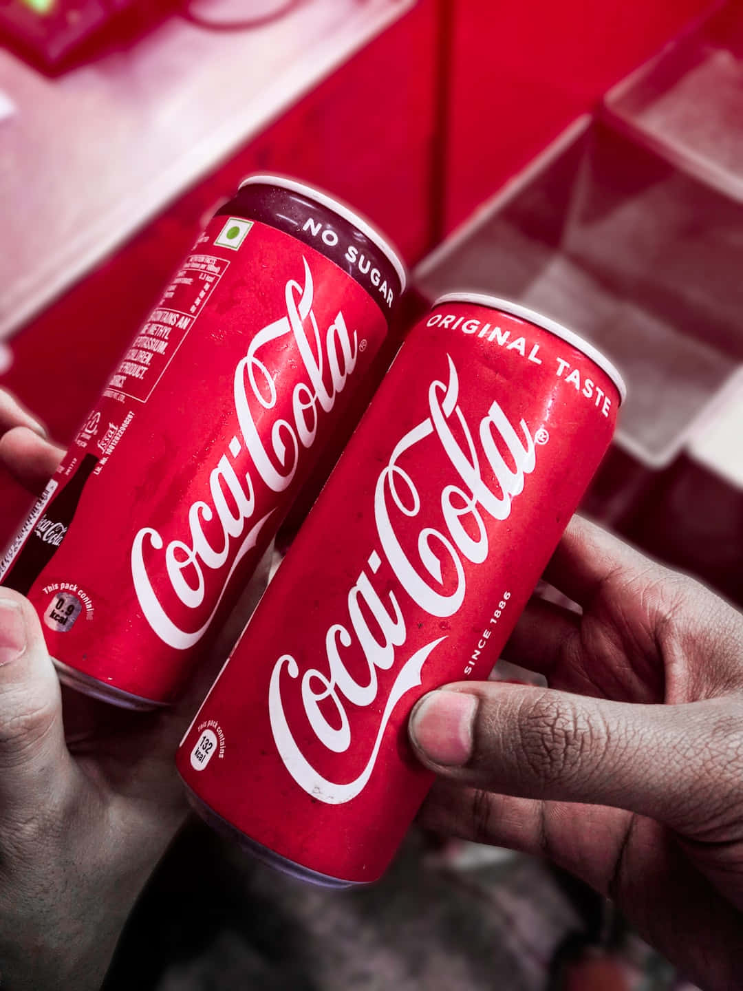coca cola 桌布 壁紙 — 兩隻手拿著紅色可口可樂罐，一個標示no sugar，另一個original taste，背景是模糊的紅色。