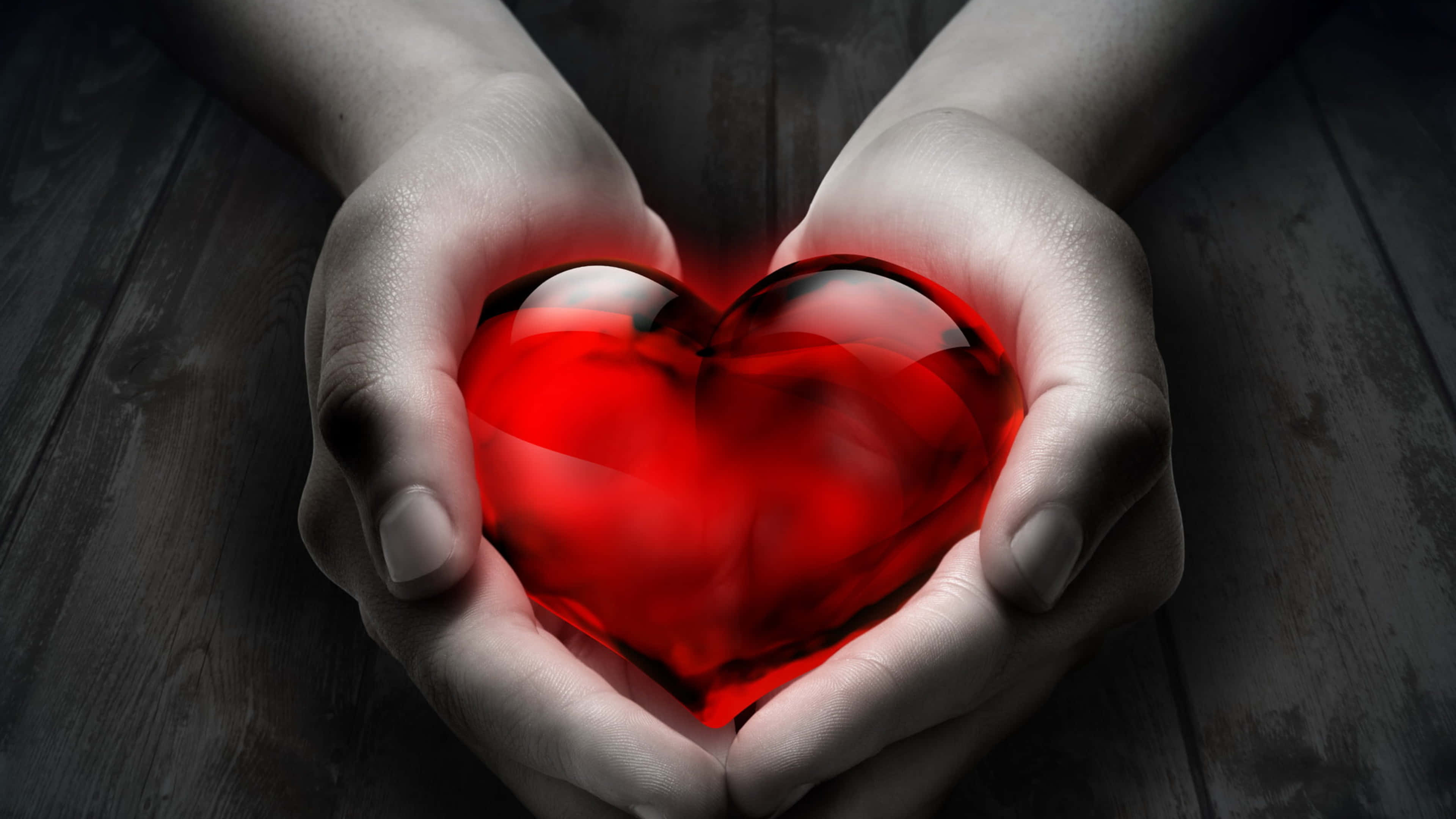 4K Love 桌布 壁紙 — 兩隻手捧著一個發光的紅色玻璃心臟，放在深色木質表面上，形成戲劇性的特寫。