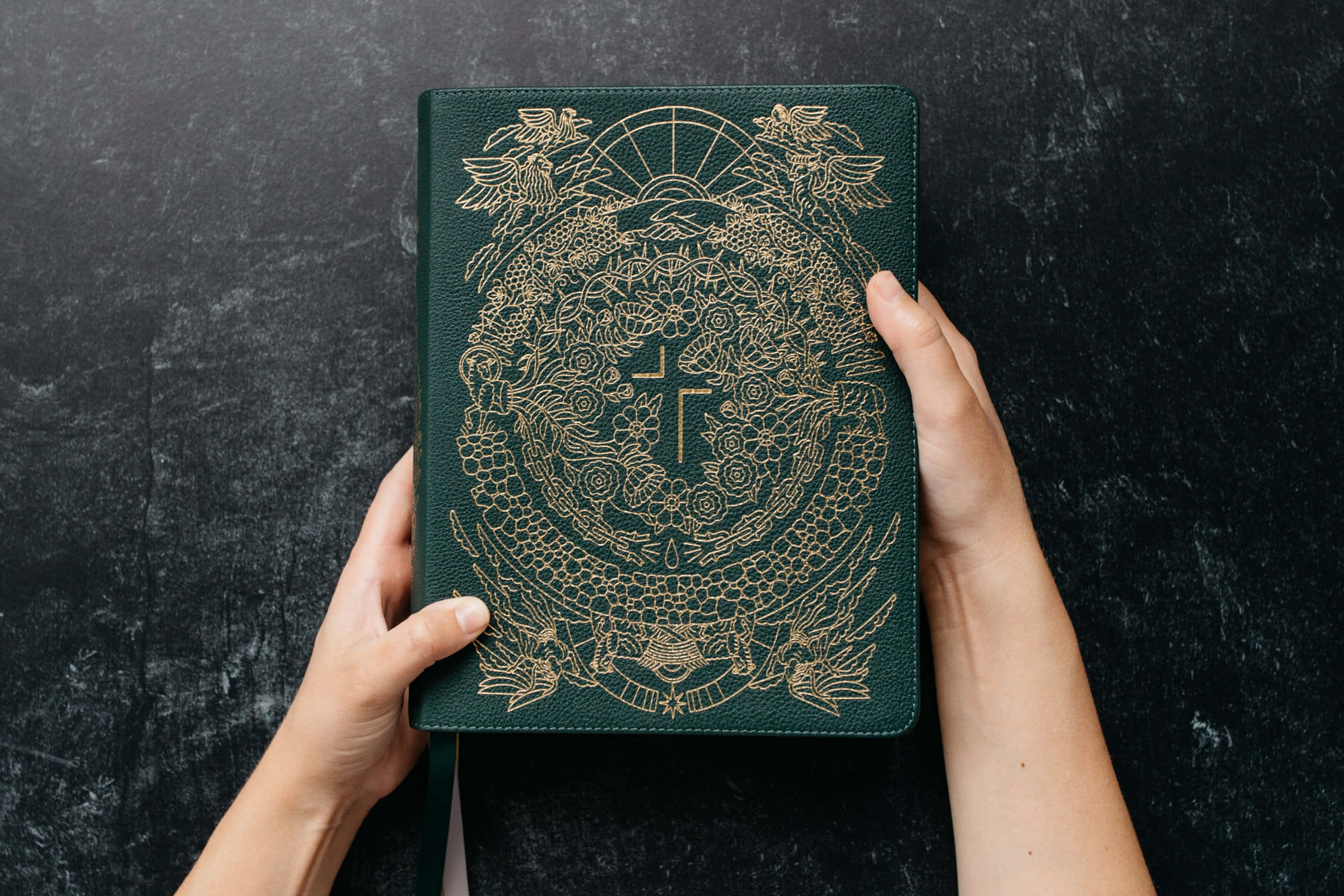 聖經 桌布 壁紙 — 兩隻手捧著一本深綠色皮革裝訂的書，書上有精緻的金色壓印龍和十字架圖案，背景是帶紋理的黑色。