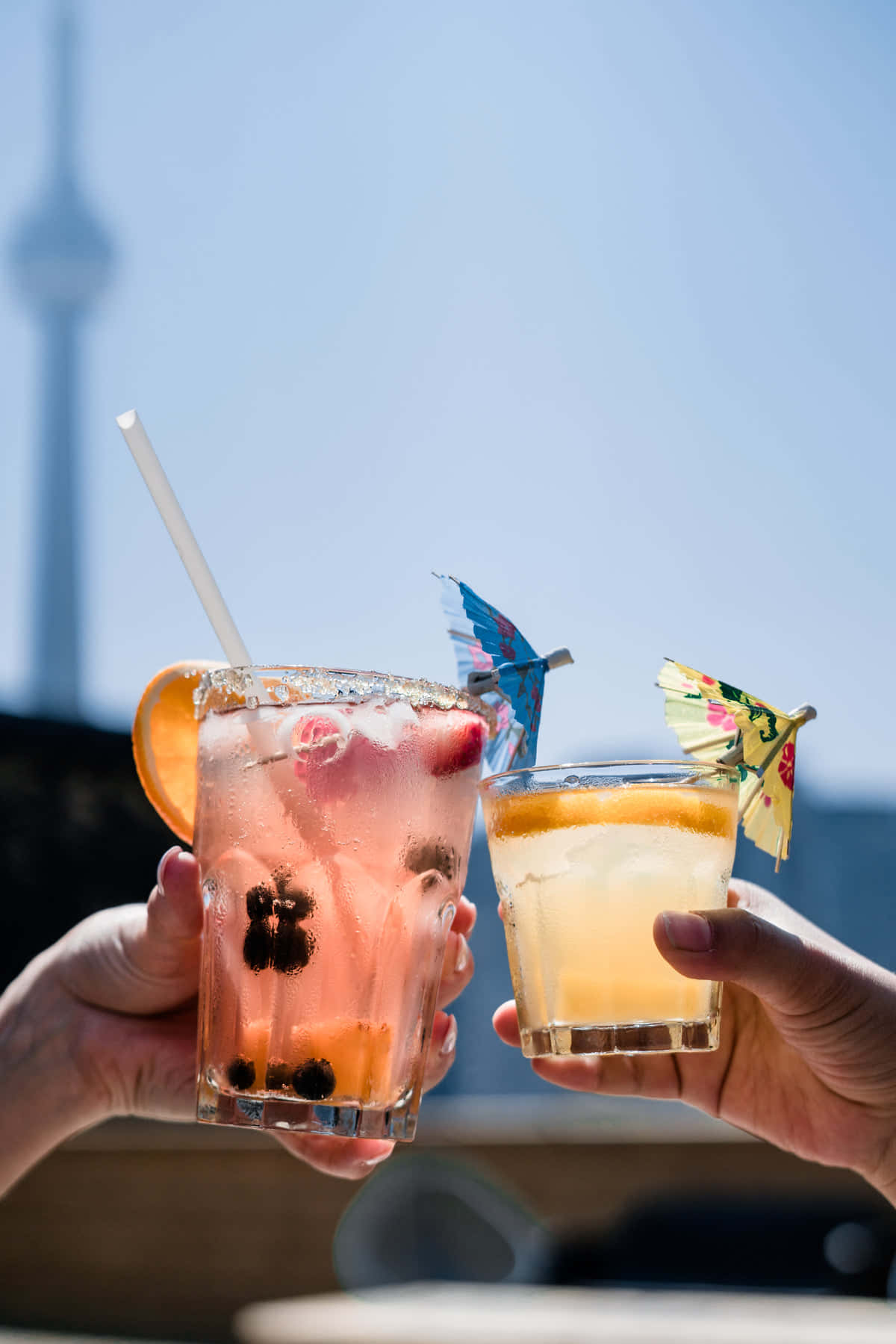 Cheers 桌布 壁紙 — 兩隻手碰杯，杯中裝有色彩繽紛的雞尾酒，飾以水果和小傘，背景是明亮的藍天和遠處的塔樓。