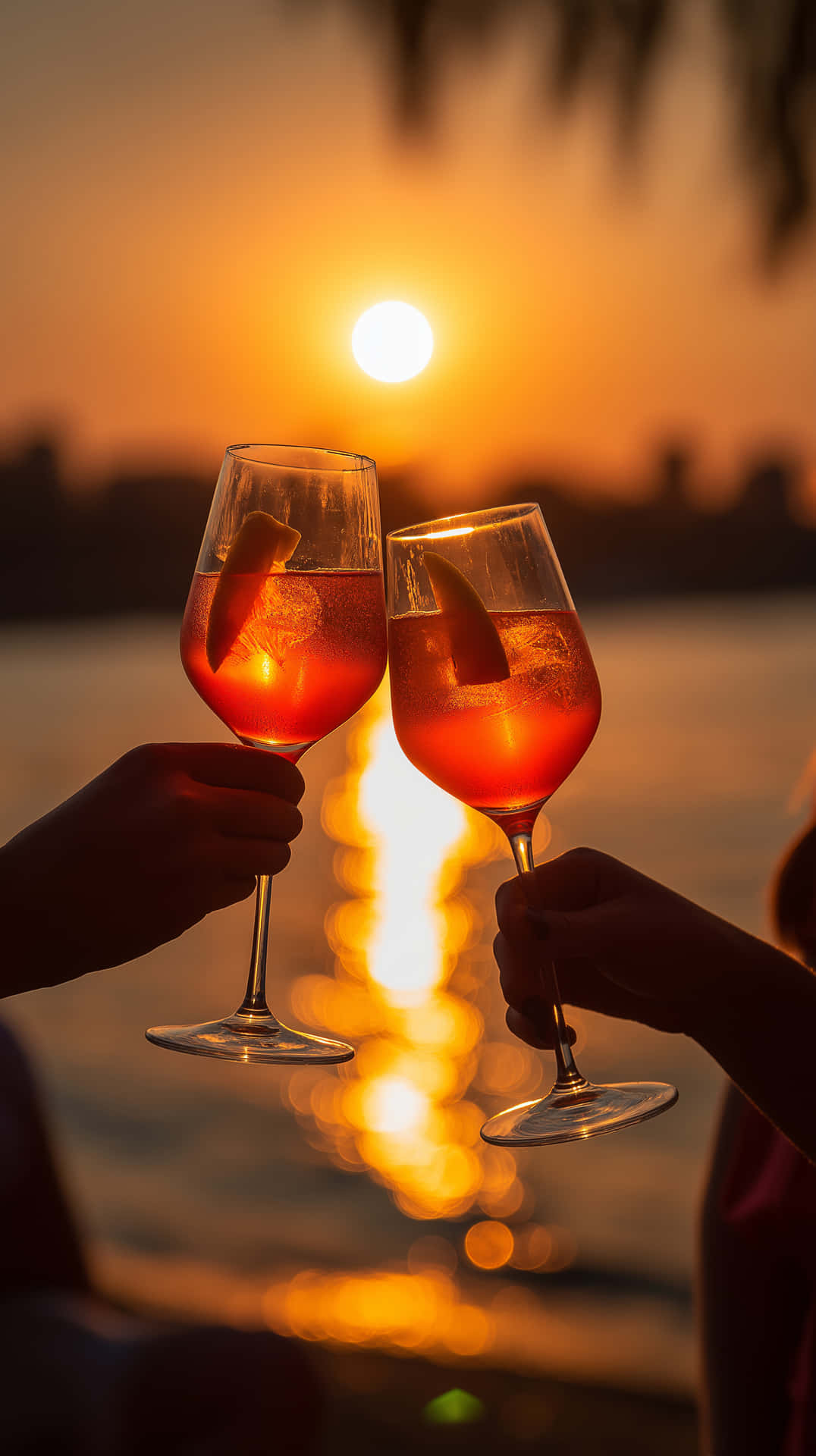Sparkling Sunset Reflections: Aperol Spritz by the Water 壁紙 — 兩隻手碰杯橙色雞尾酒，背景是水面上的夕陽餘暉，上方有棕櫚葉的剪影。