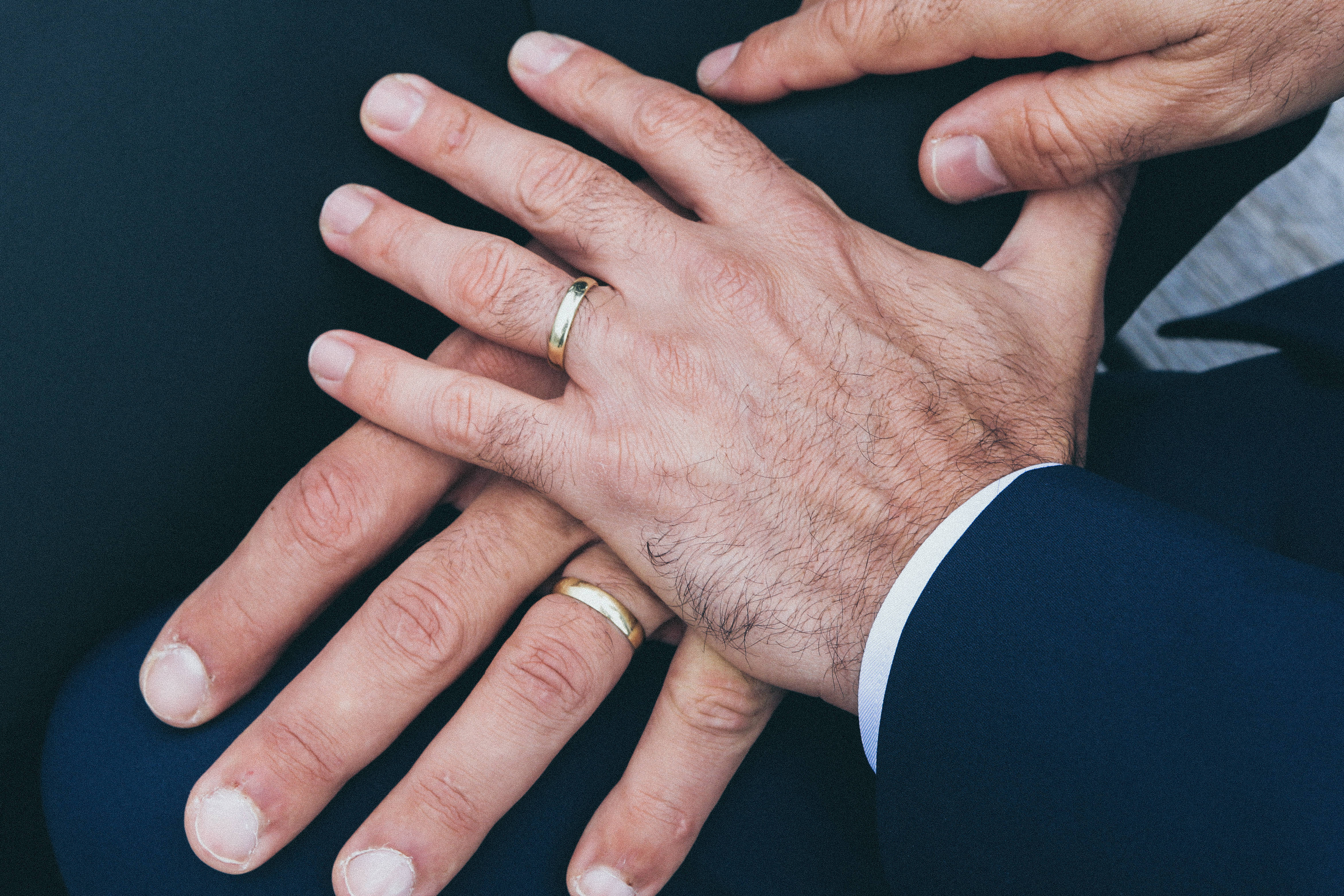 男同志 桌布 壁紙 — 兩隻手緊握在一起，一隻戴著結婚戒指，另一隻戴著簡單的金戒指，背景為深色布料。
