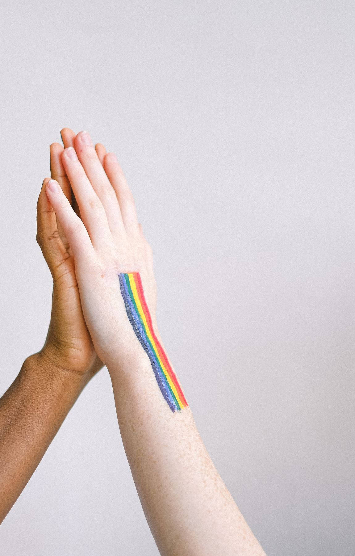 Subtle Lgbt 桌布 壁紙 — 兩隻手緊握在一起，其中一隻手腕上有鮮豔的彩虹旗刺青，背景為純色。