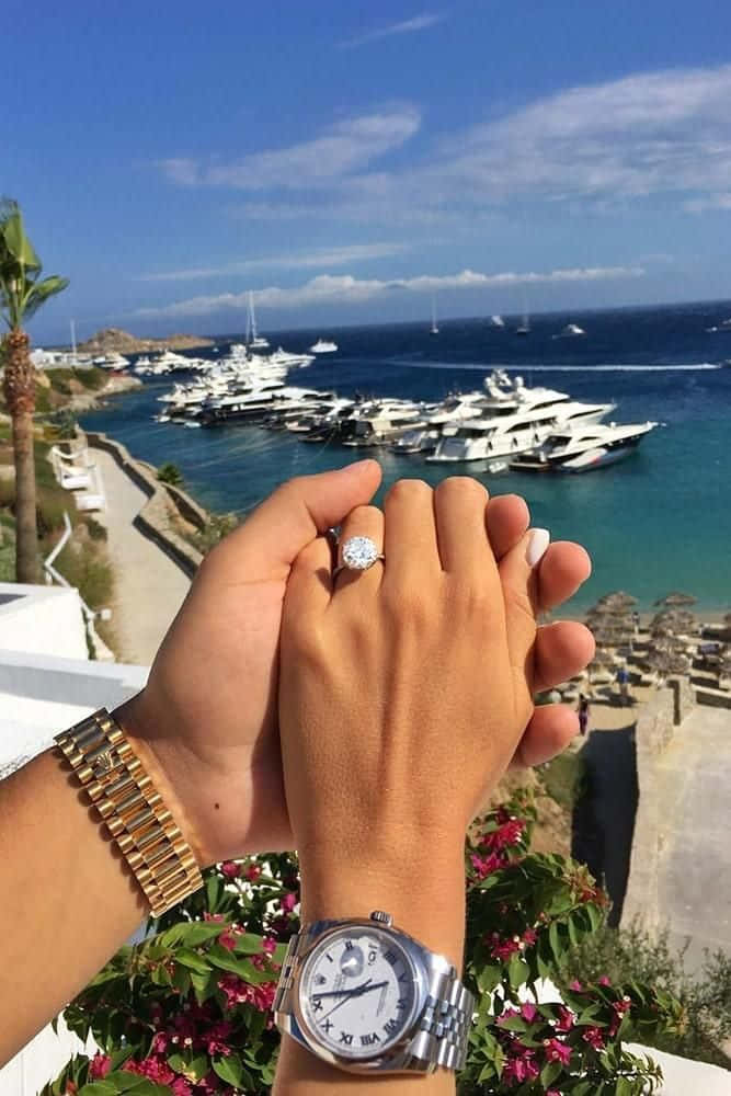 engagement 桌布 壁紙 — 兩隻手緊握在一起，展示閃閃發光的訂婚戒指和奢華手錶，背景是充滿遊艇和碧綠海水的碼頭。