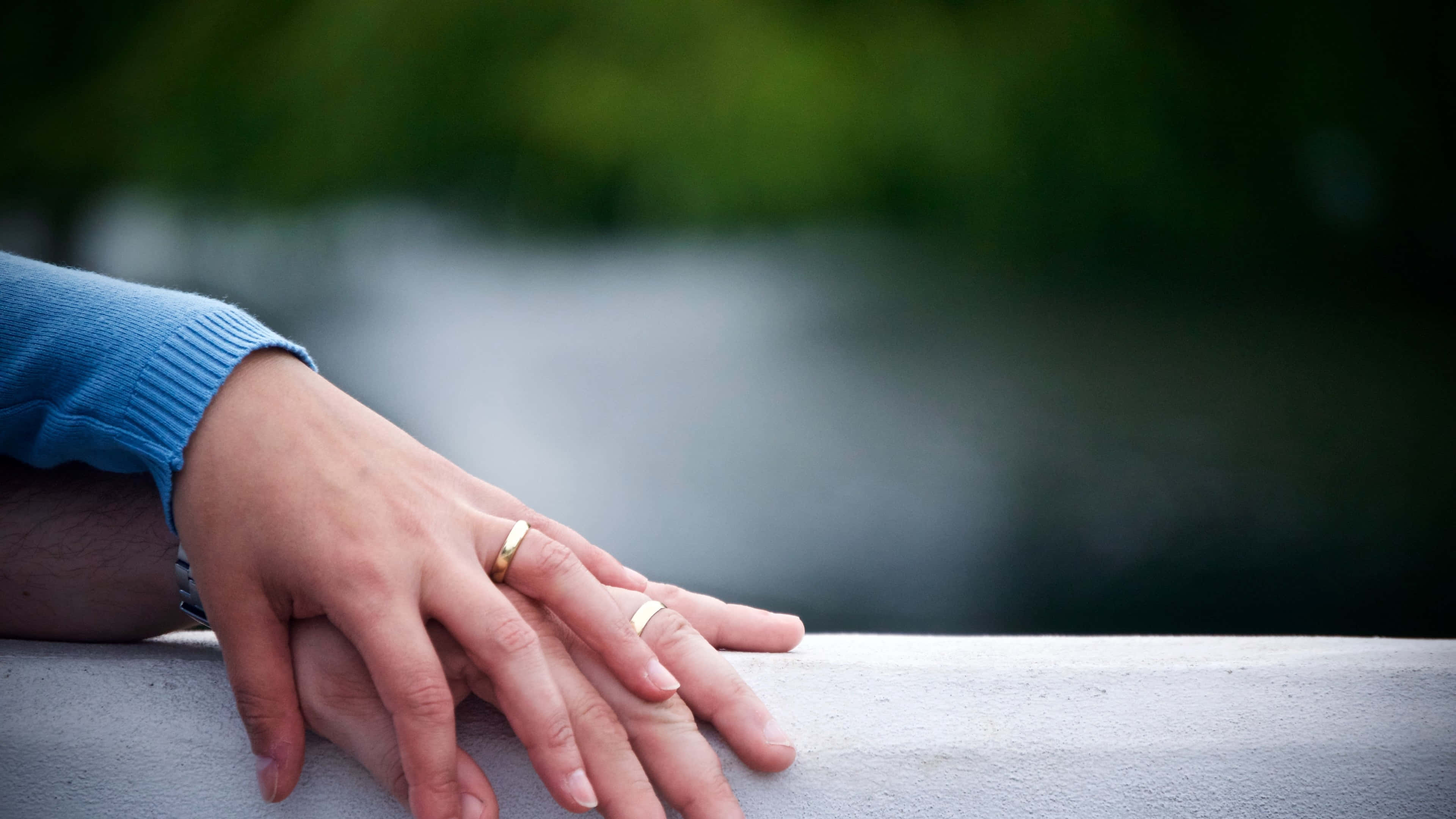 Wedding Rings 桌布 壁紙 — 兩隻手緊握在一起，戴著結婚戒指，擱在石頭邊緣上，背景是模糊的綠色和灰色。