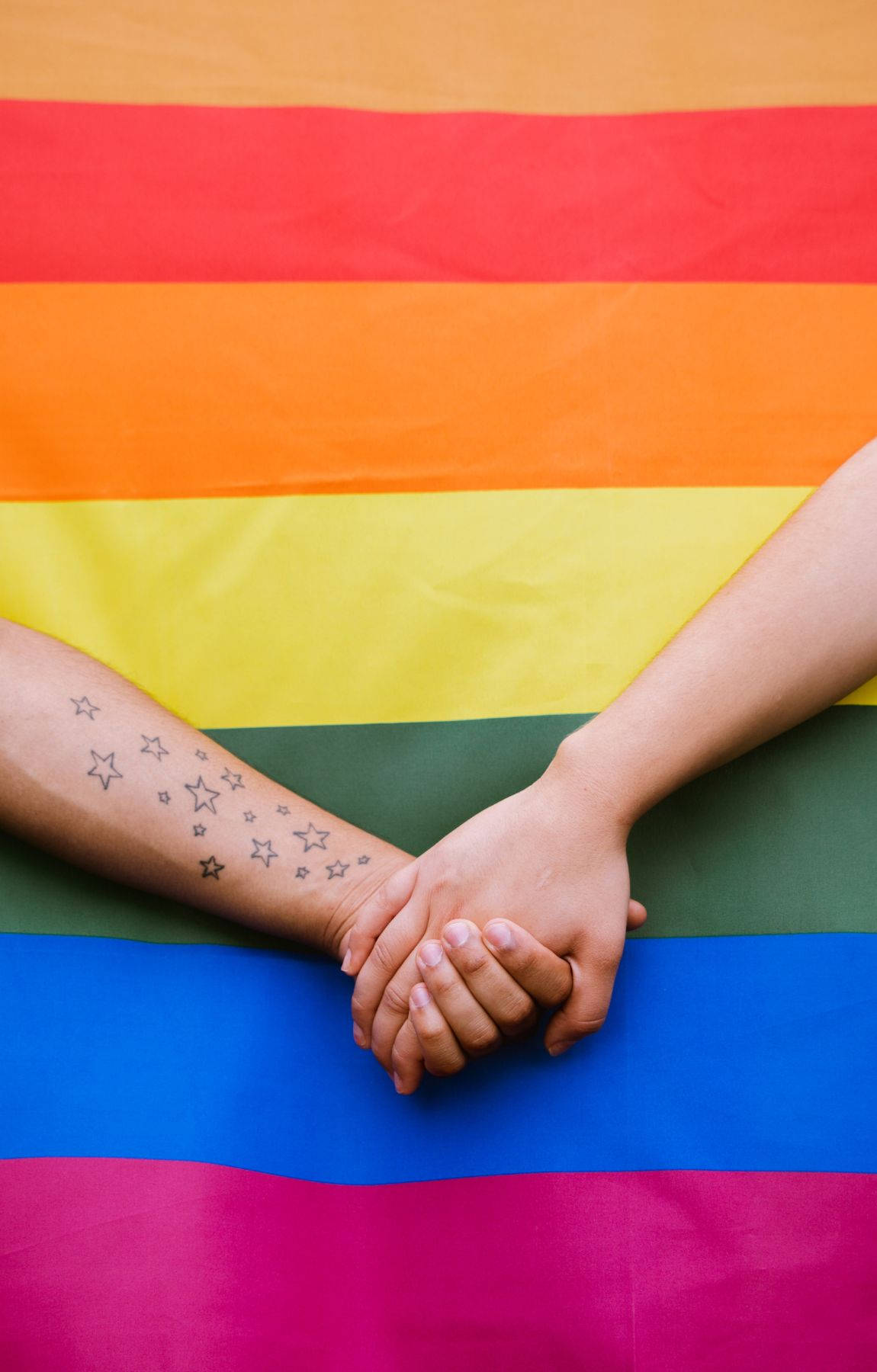 Subtle Lgbt 桌布 壁紙 — 兩隻手緊握在一起，置於鮮豔的彩虹旗之上，其中一隻手臂上有星星刺青。
