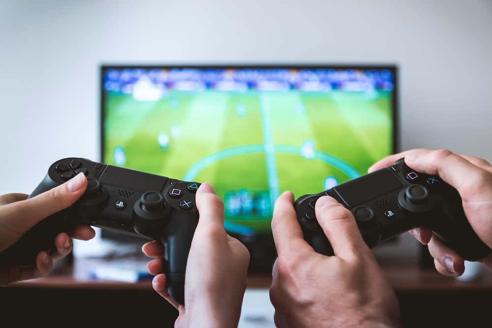 PlayStation 桌布 壁紙 — 兩隻手緊握黑色遊戲控制器，專注於電視螢幕上顯示的一場充滿活力的足球比賽。
