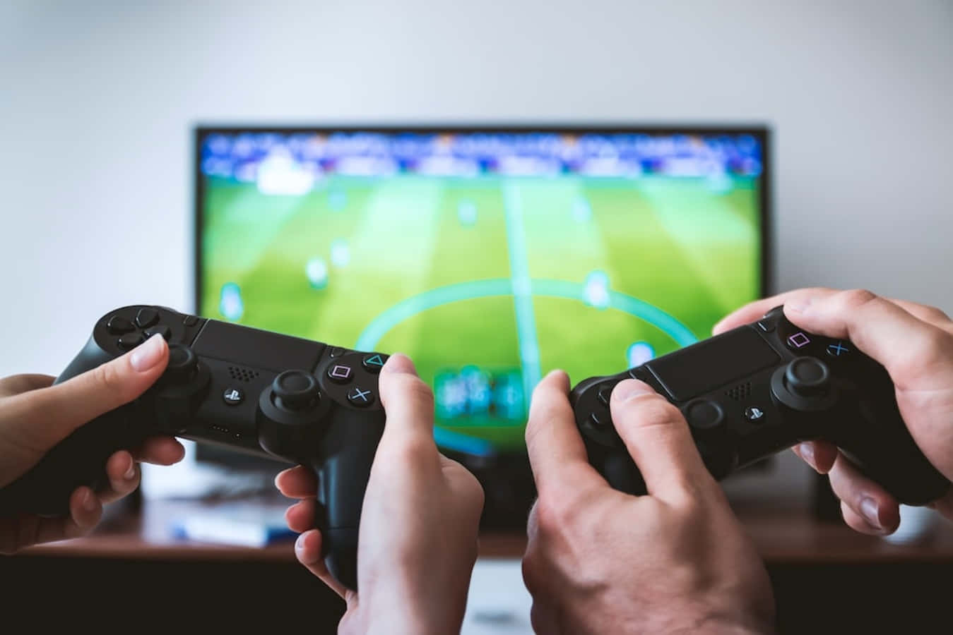 game 桌布 壁紙 — 兩隻手緊握黑色遊戲控制器，專注於電視螢幕上顯示的一場充滿活力的足球比賽。