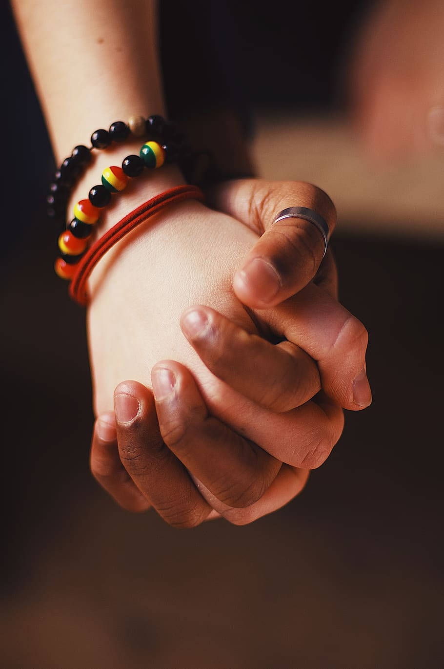 hands 桌布 壁紙 — 兩隻手緊緊相握，一隻戴著珠串手鍊和戒指，另一隻裸露，背景是暗色模糊的。