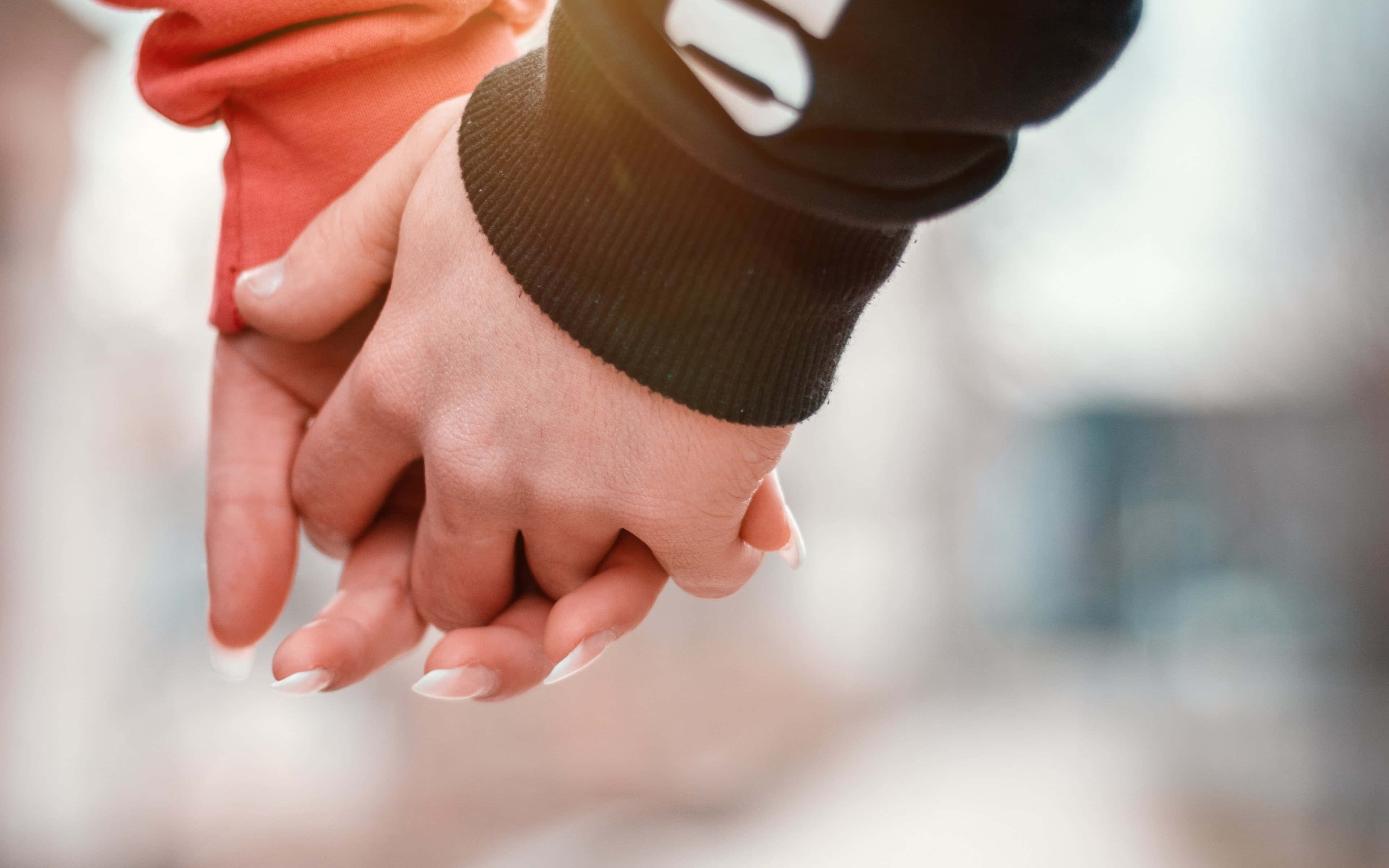 Holding Hands 桌布 壁紙 — 兩隻手緊緊相握，一隻穿著黑色袖子，另一隻穿著橙色袖子並塗有白色指甲油，背景柔和模糊。