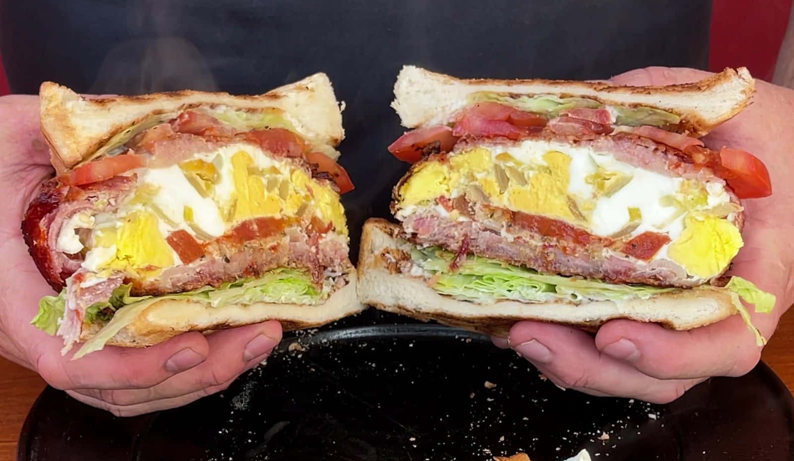 bacon 桌布 壁紙 — 兩隻手自豪地展示一個新鮮切開的培根蛋起司三明治，露出融化起司、火腿、番茄和生菜的層次。