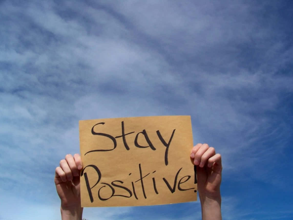 正向 桌布 壁紙 — 兩隻手舉起一個手製紙板標語，上面寫著「Stay Positive!」，背景是晴朗的藍天和縷縷白雲。