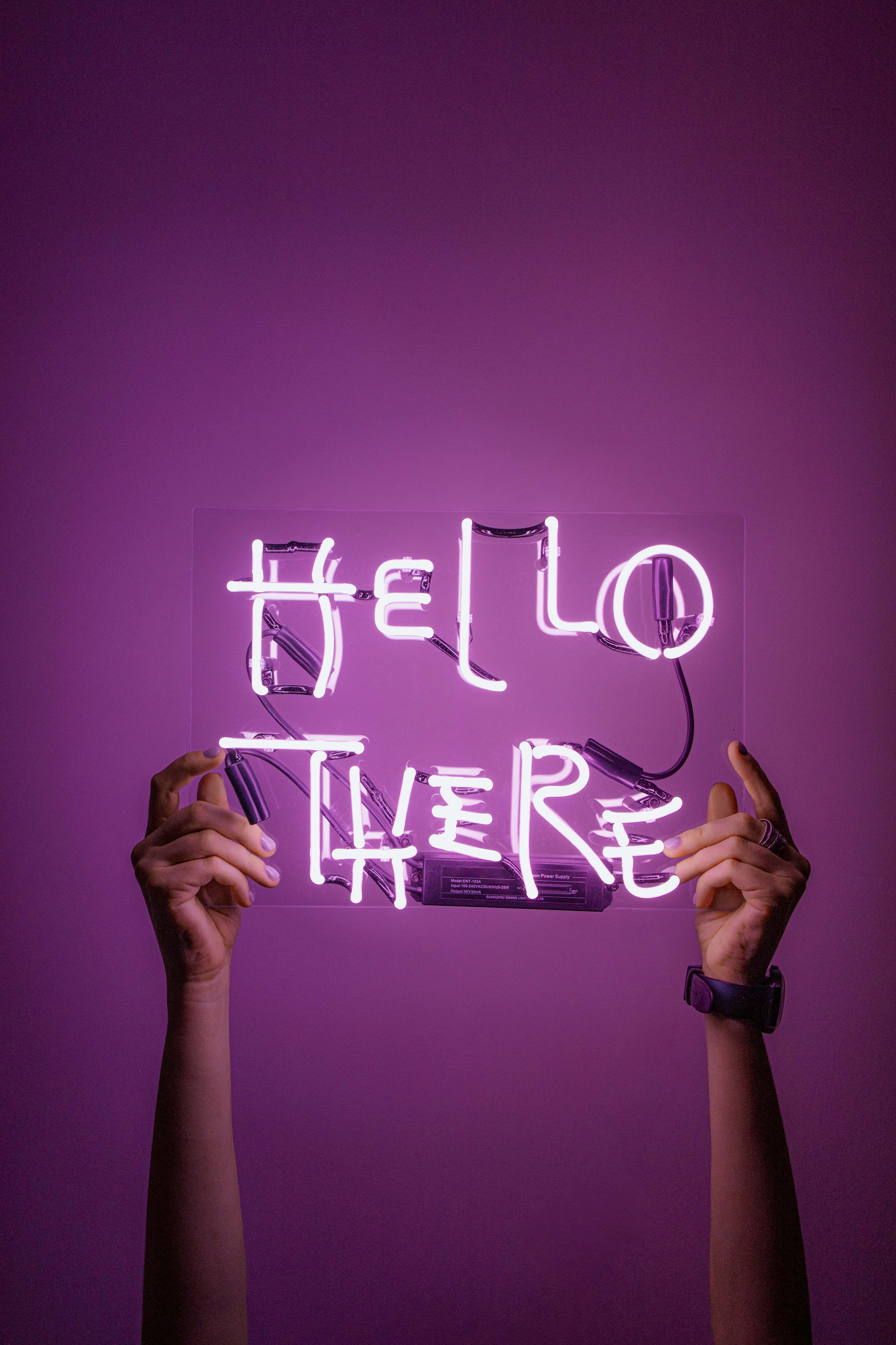 Hello 桌布 壁紙 — 兩隻手舉起一個發光的霓虹燈牌，上面寫著 hello there，背景是純紫色牆壁。