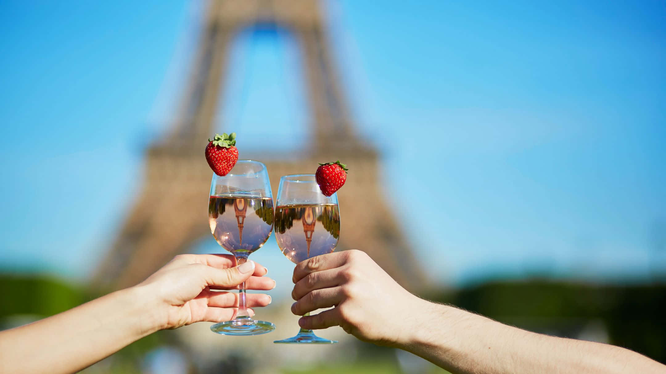 Eiffel Tower 桌布 壁紙 — 兩隻手舉起帶有草莓的玫瑰酒杯碰杯，背景是模糊的艾菲爾鐵塔，在明亮的藍天下。