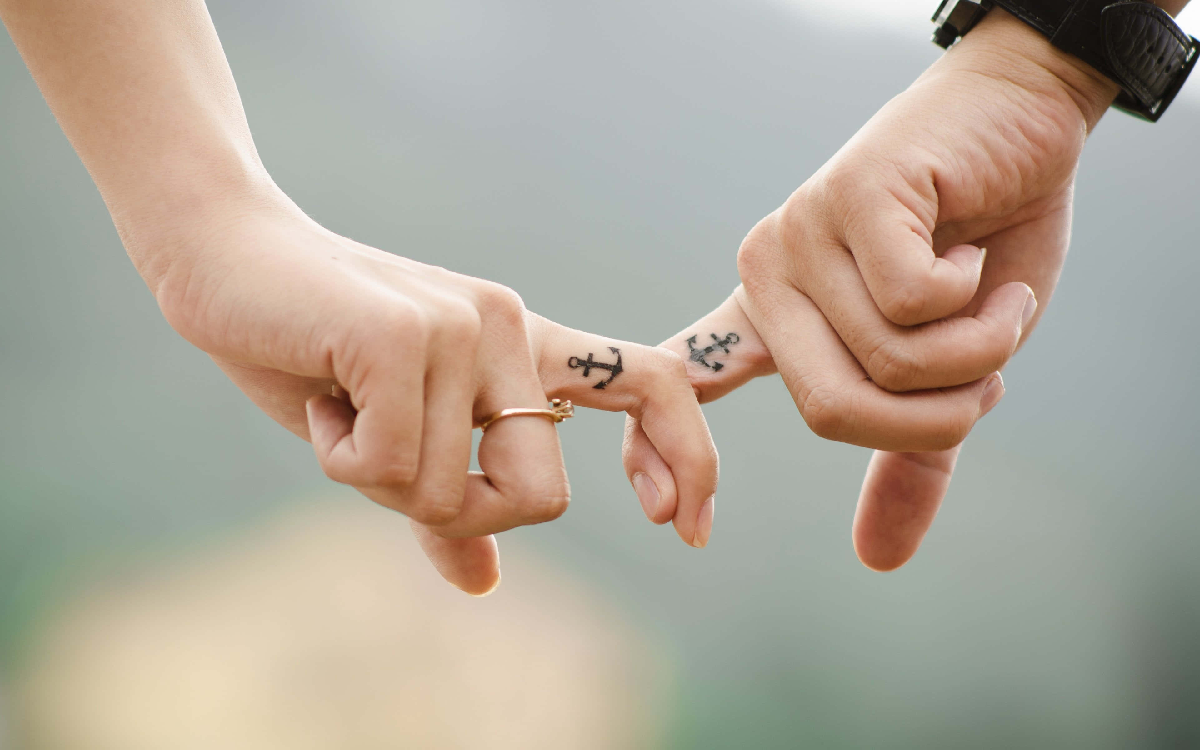 anchors 桌布 壁紙 — 兩隻手輕輕交握，指尖有相配的錨形刺青，背景是柔和模糊的自然景色。