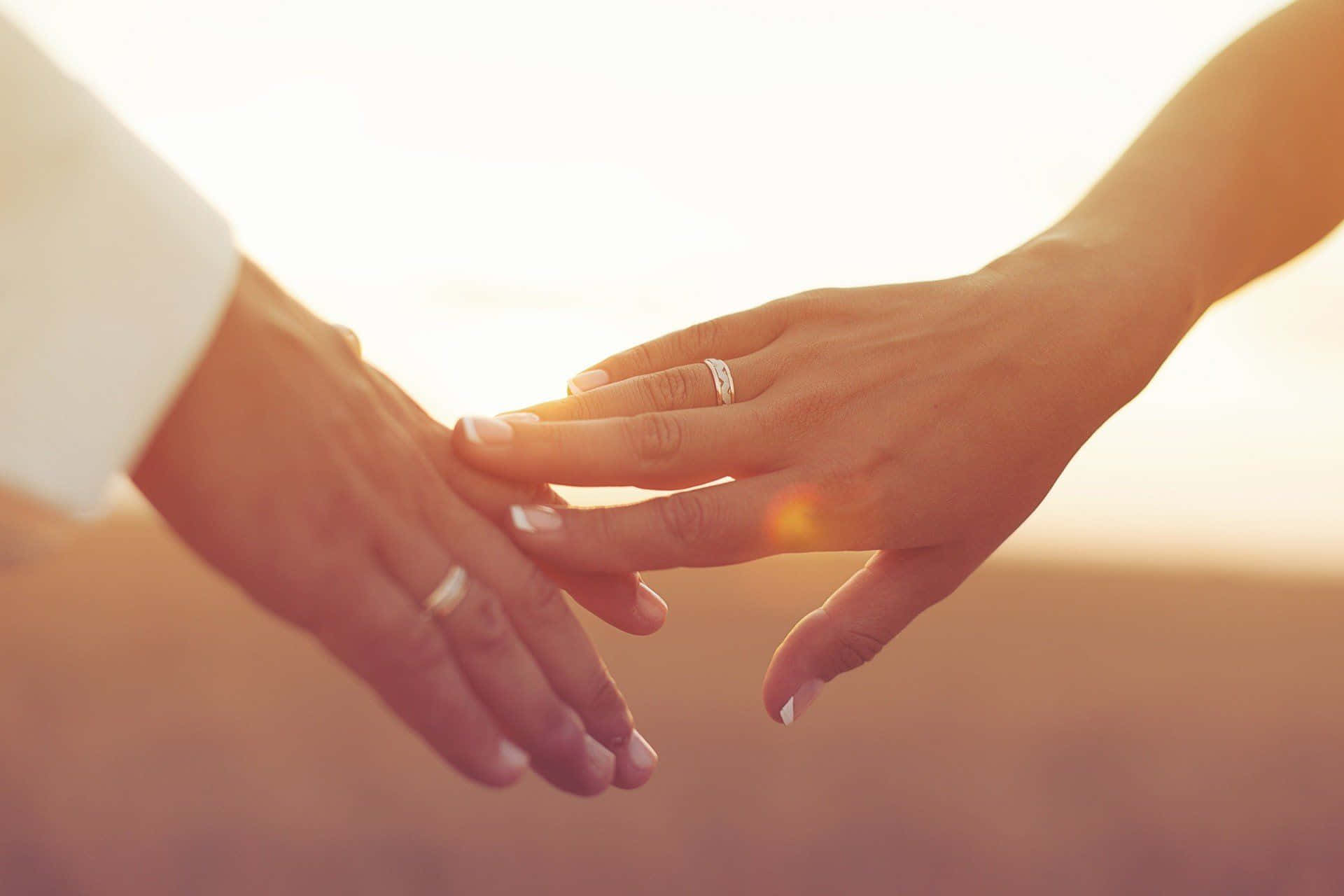 日落 桌布 壁紙 — 兩隻手輕輕交握，結婚戒指在溫暖的金色夕陽光中閃耀。