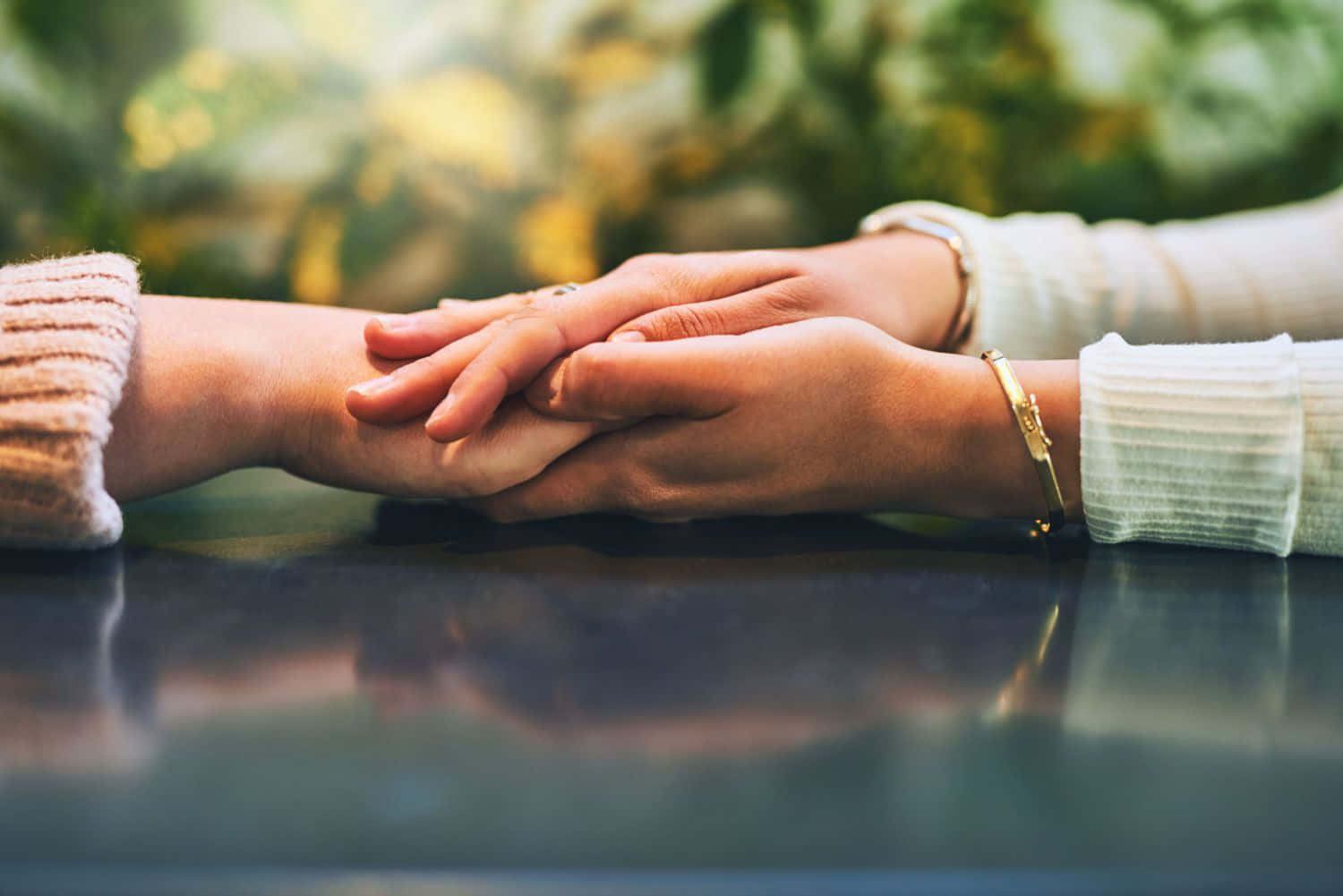 forgive 桌布 壁紙 — 兩隻手輕輕交握在反光表面上，背景是柔和模糊的綠色植物。