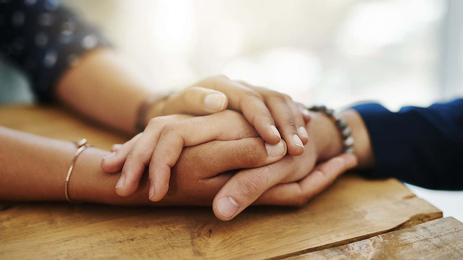 Kind 桌布 壁紙 — 兩隻手輕輕交握在溫暖的木質表面上，傳達舒適與連結。