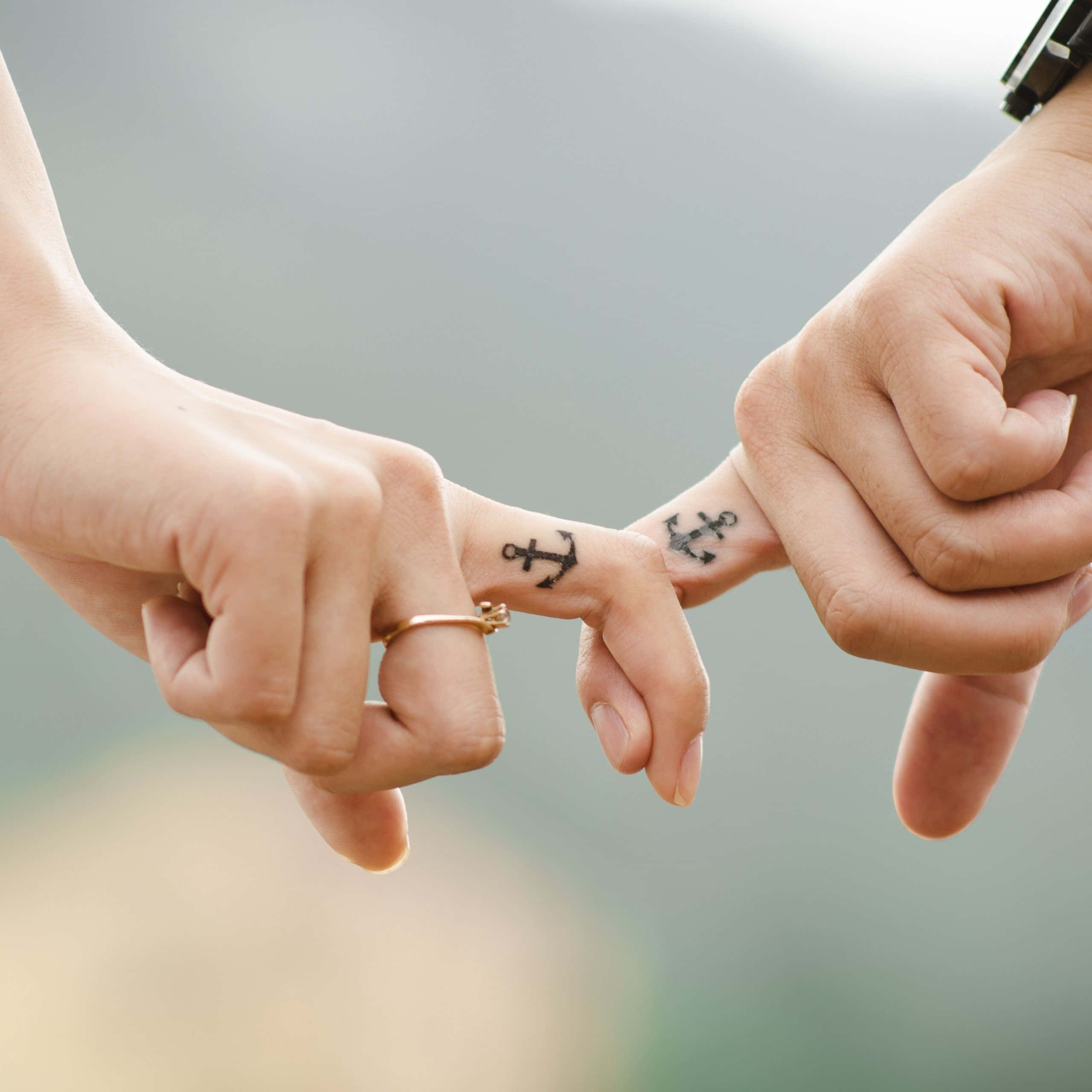 couple love 桌布 壁紙 — 兩隻手輕輕觸碰指尖，露出相配的錨紋身，象徵著在柔和模糊背景下的牢固聯繫。