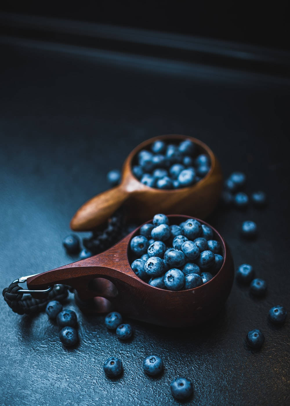 Blueberry 桌布 壁紙 — 兩隻木匙盛滿飽滿的藍莓，放在深色紋理表面上，周圍散落著莓果。