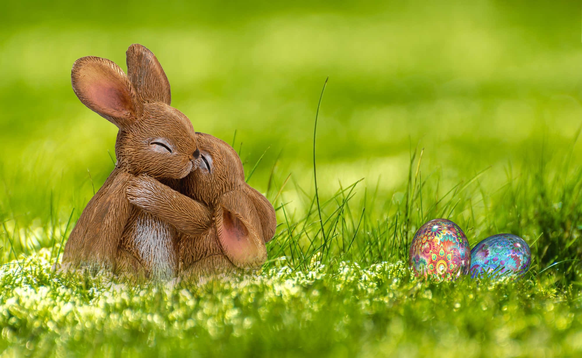 Easter 桌布 壁紙 — 兩隻棕色兔子公仔在鮮綠色草地上，在色彩繽紛的圖案復活蛋旁邊溫柔擁抱。