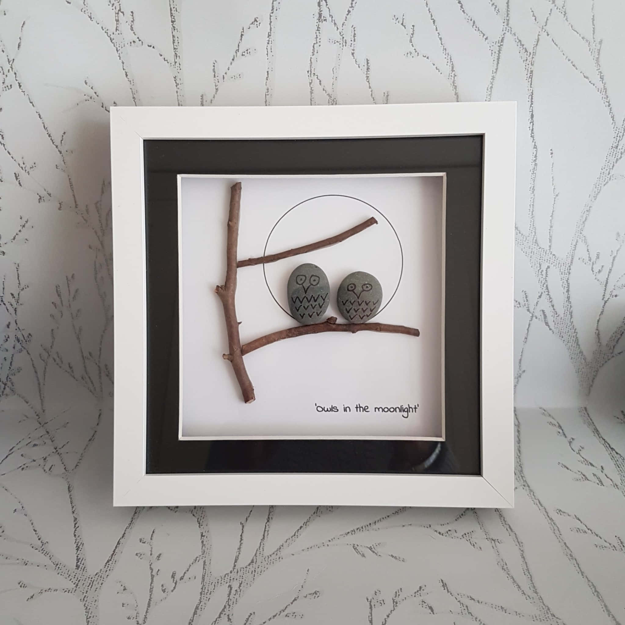 Pebble Art 桌布 壁紙 — 兩隻灰色石頭貓頭鷹坐在樹枝上，下方有繪製的月亮，置於白色陰影框中，背景為樹紋牆壁。