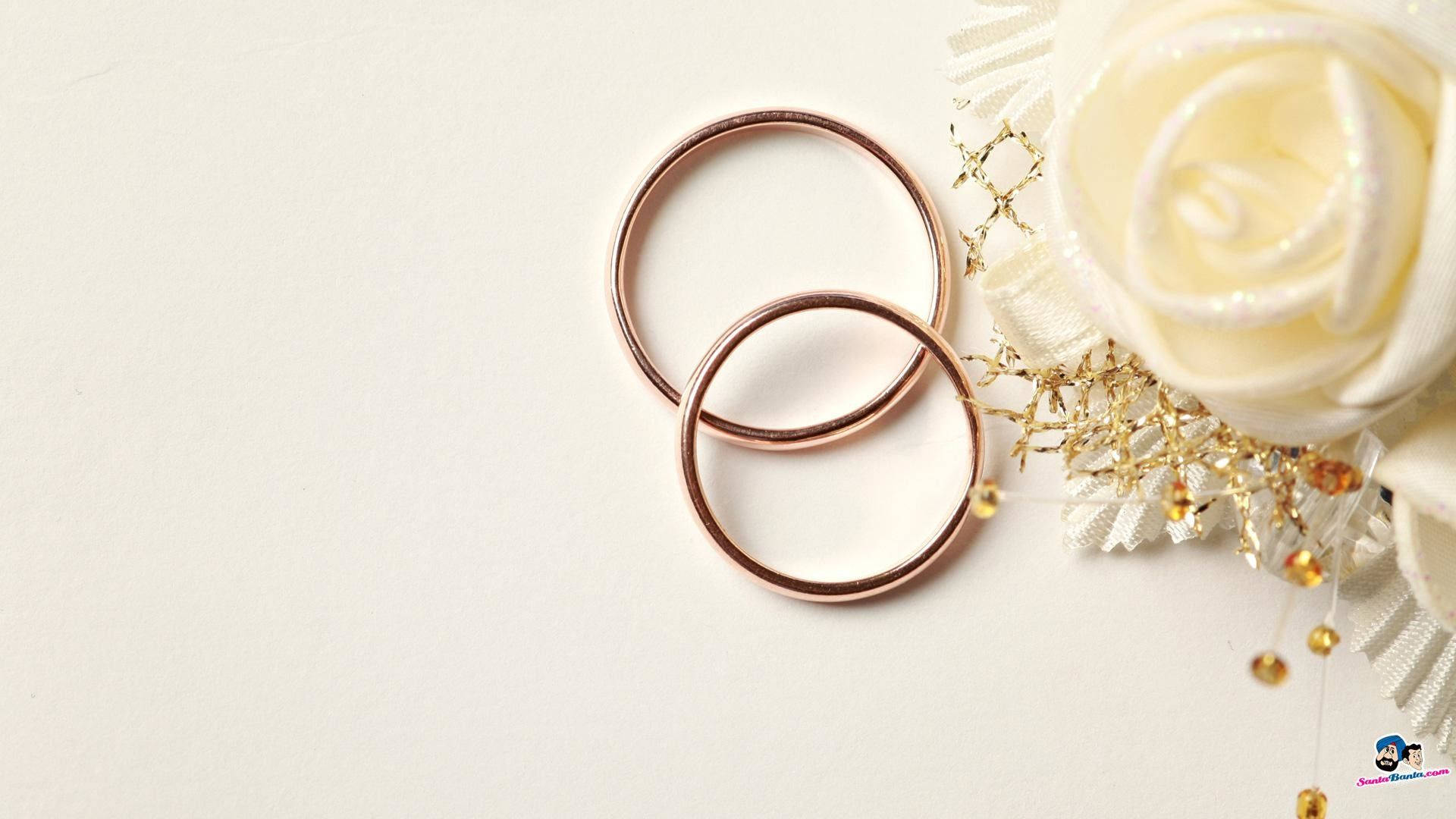 Wedding Rings 桌布 壁紙 — 兩隻玫瑰金結婚戒指放在白色玫瑰和金飾旁邊，置於潔白表面上。