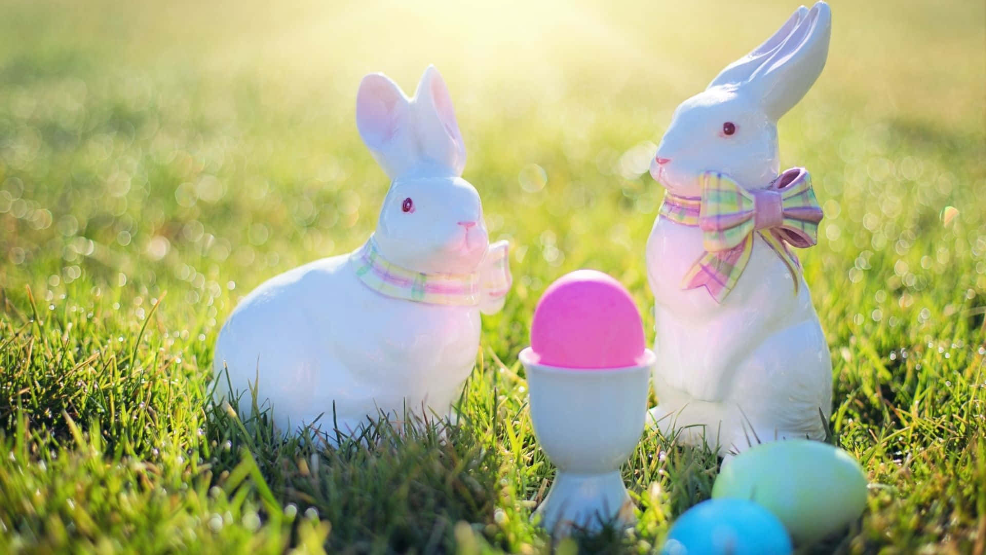 Easter Bunny 桌布 壁紙 — 兩隻白色陶瓷復活節兔子繫著粉彩蝴蝶結，坐在陽光普照的草地上，旁邊是色彩繽紛的蛋，構成明亮的春天場景。
