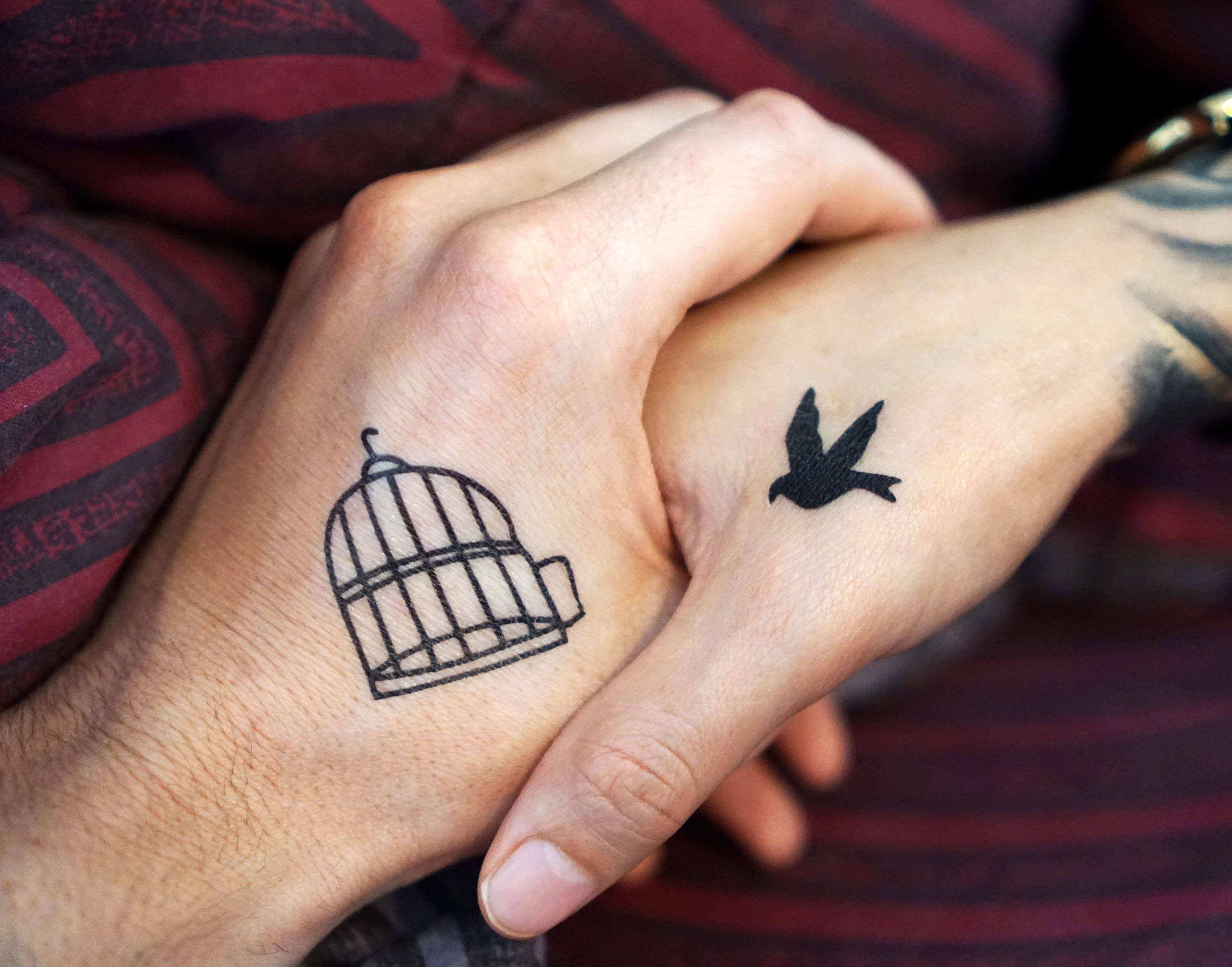 tattoos 桌布 壁紙 — 兩隻緊握的手，展示鳥籠和一隻飛鳥的刺青，象徵自由與連結。