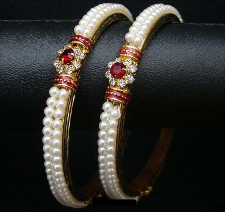 Bangles 桌布 壁紙 — 兩隻華麗的金色手鐲，飾有白色珍珠和紅色寶石點綴，優雅地放置在黑色皮革表面上。