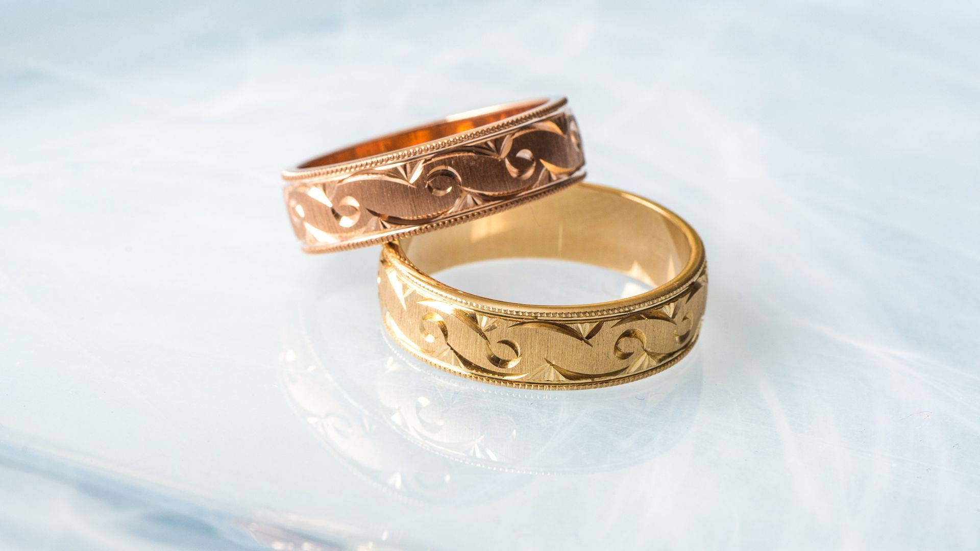 Wedding Rings 桌布 壁紙 — 兩隻華麗的金色結婚戒指，帶有複雜的捲曲花紋，放置在光滑的反射表面上。