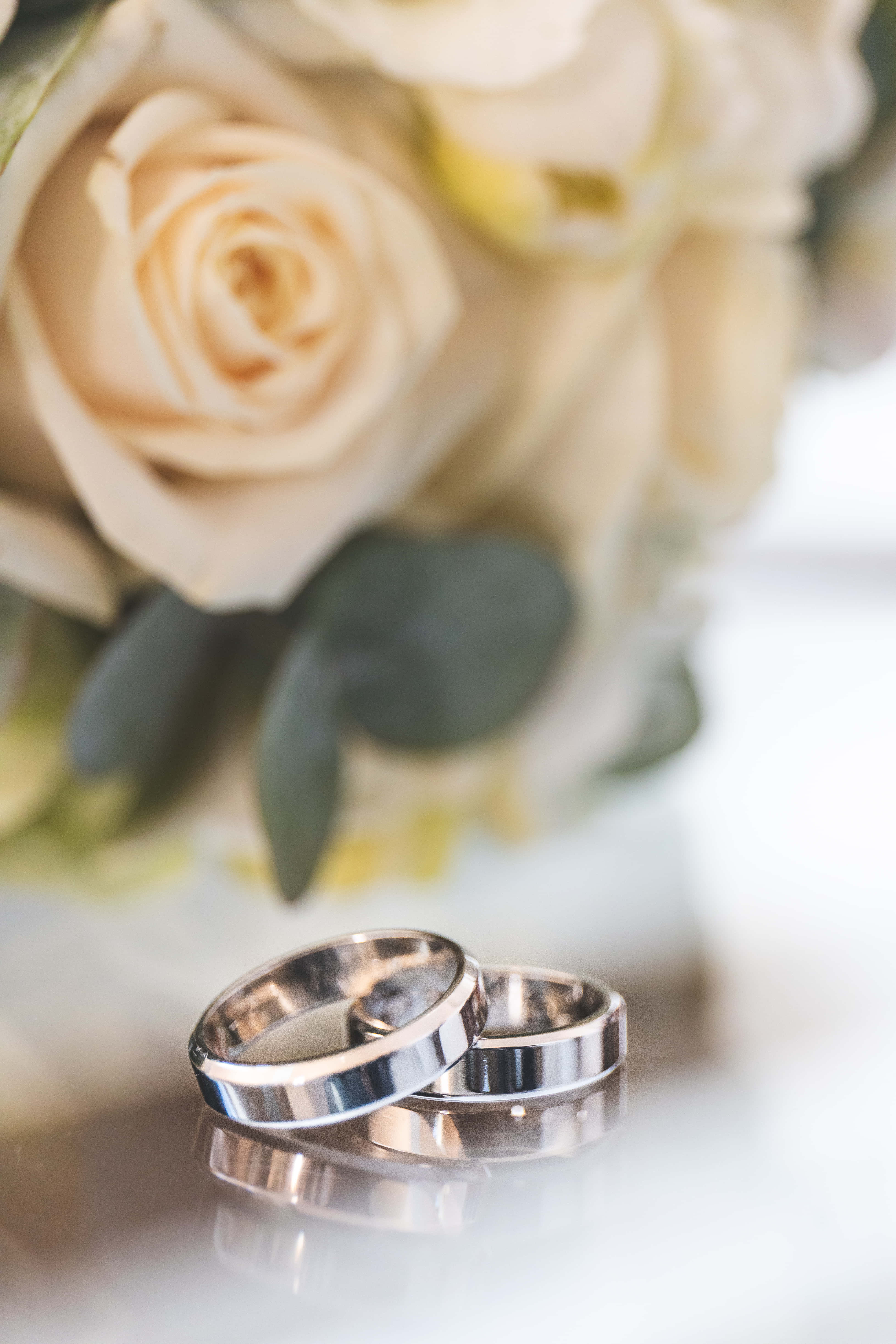 Marriage 桌布 壁紙 — 兩隻閃亮的結婚戒指放在反光表面上，後面有一束柔焦的奶油色玫瑰和綠葉。