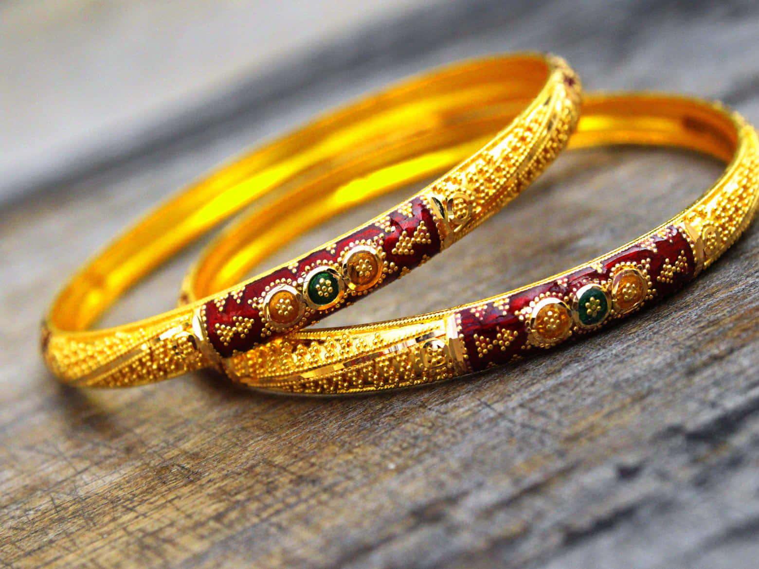 Bangles 桌布 壁紙 — 兩隻飾有紅色琺瑯和綠色寶石的華麗金手鐲，放在經風化的木質表面上。
