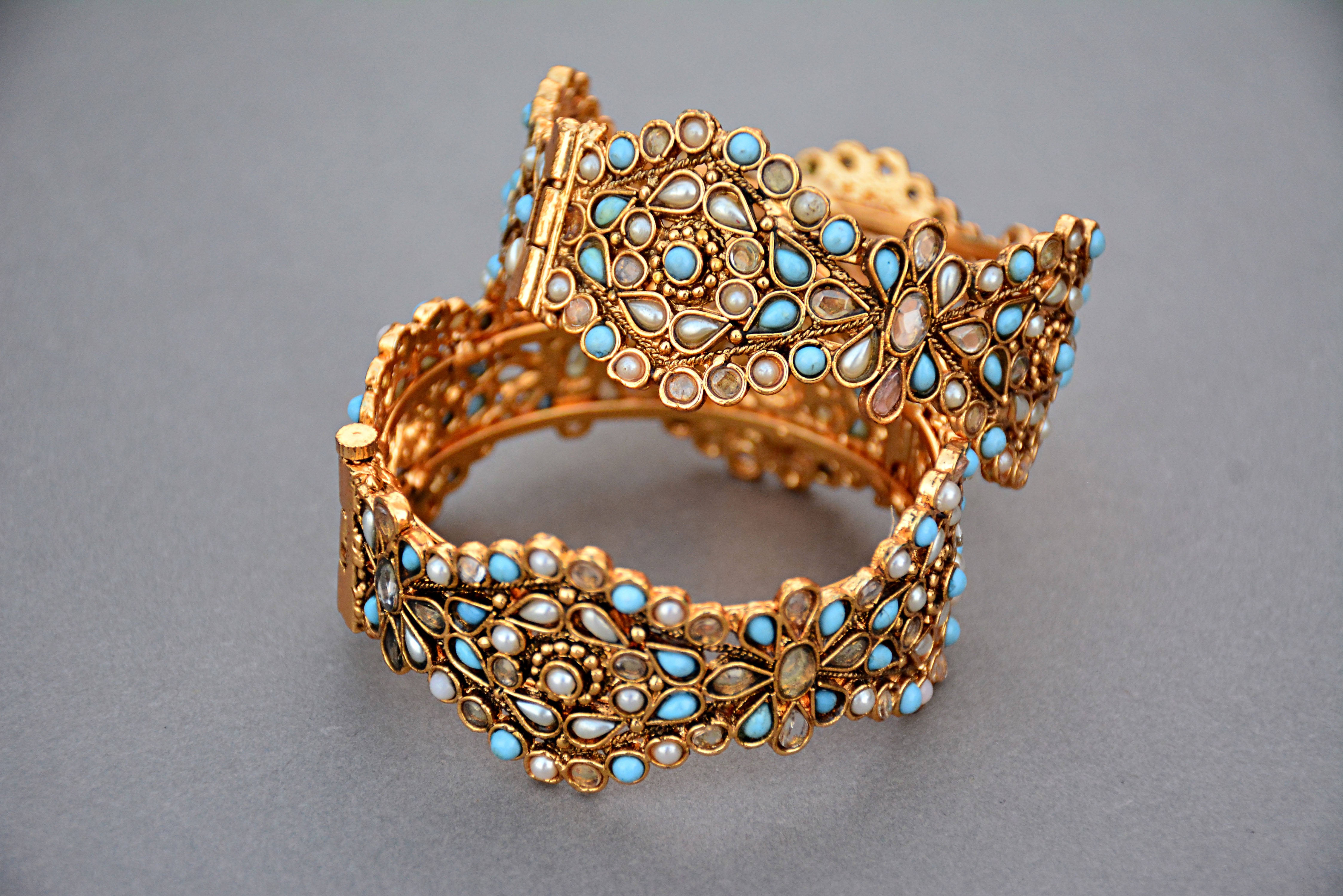 Gold Jewellery 桌布 壁紙 — 兩隻飾有綠松石和珍珠鑲嵌花卉圖案的華麗金手鐲，放置在中性灰色表面上。
