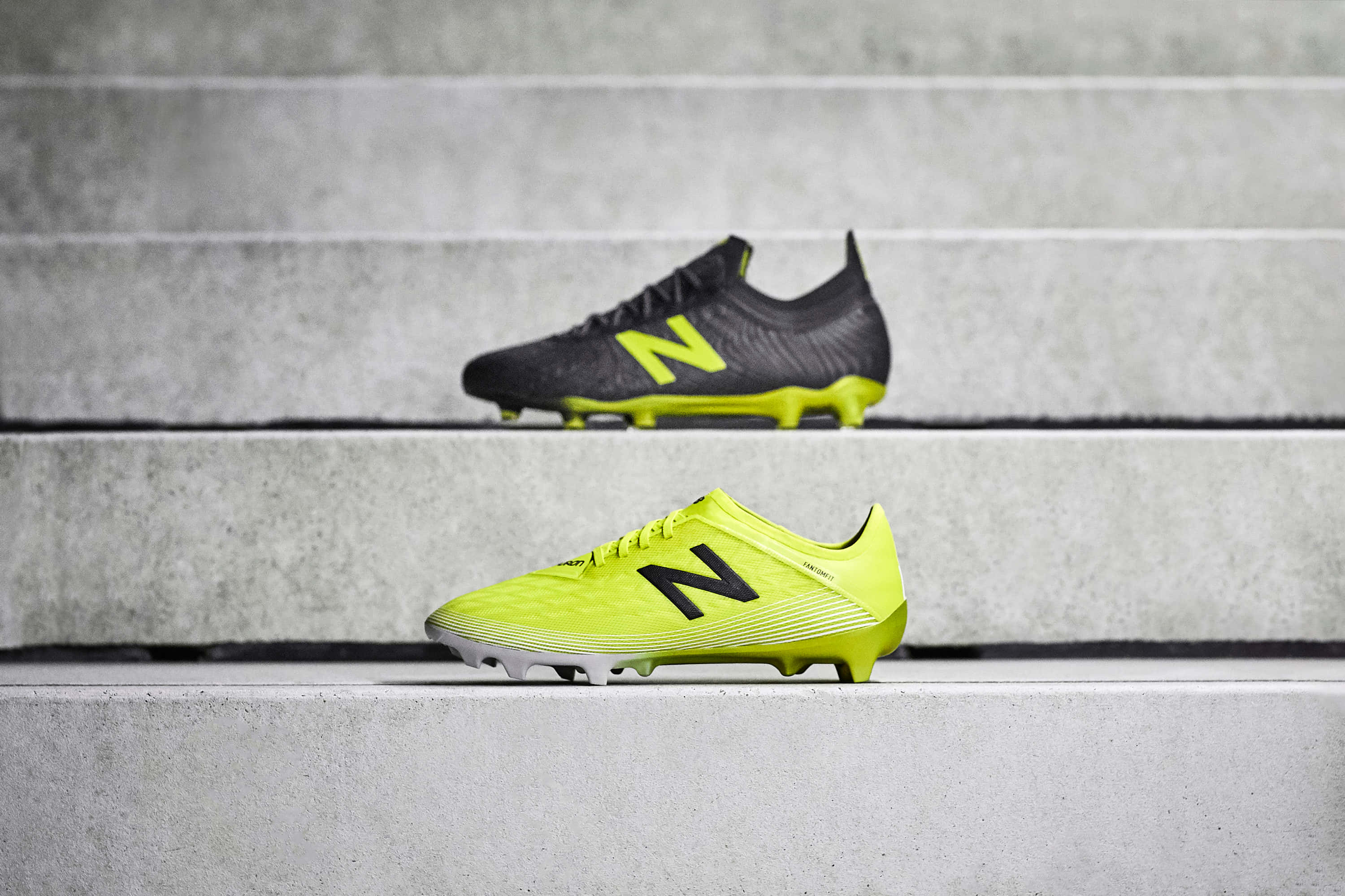 New Balance 桌布 壁紙 — 兩雙New Balance足球鞋，一雙螢光黃色，一雙黑色，放在灰色混凝土台階上。