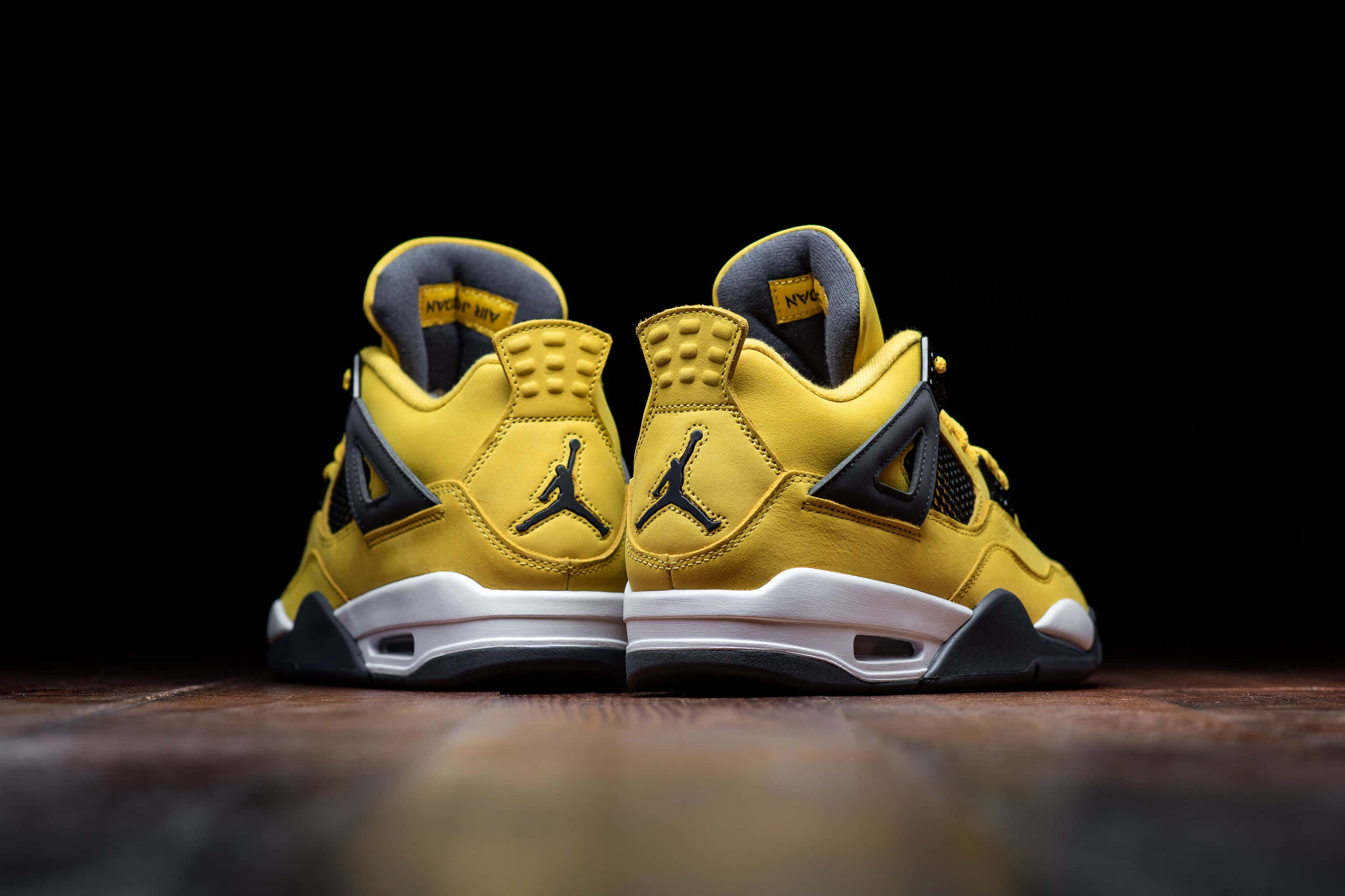 Yellow Jordan 桌布 壁紙 — 兩雙大膽的黃色Jordan 4運動鞋帶有黑色點綴，背對背放在木地板上，背景為黑色。