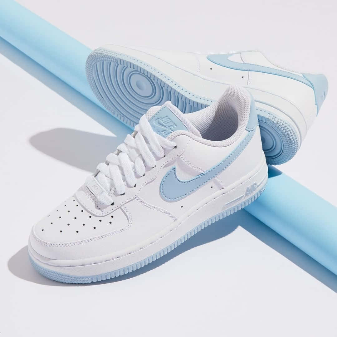Nike Air Force 桌布 壁紙 — 兩雙帶有淺藍色點綴的白色Nike Air Force 1運動鞋，放置在一個相配的藍色圓柱形物體上，背景為明亮的白色。