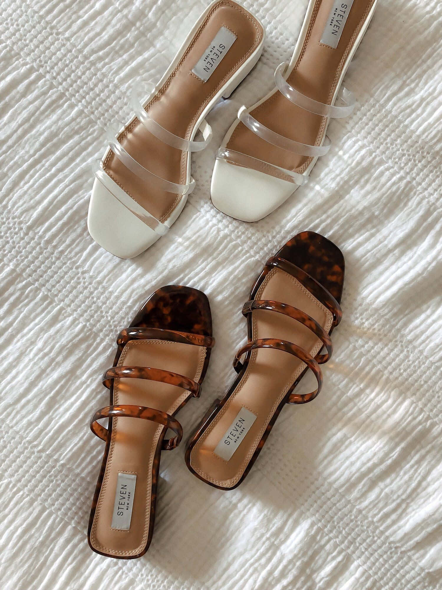 Sandals 桌布 壁紙 — 兩雙時尚的方頭涼鞋，搭配透明帶子，放置在帶有紋理的白色布料表面上。