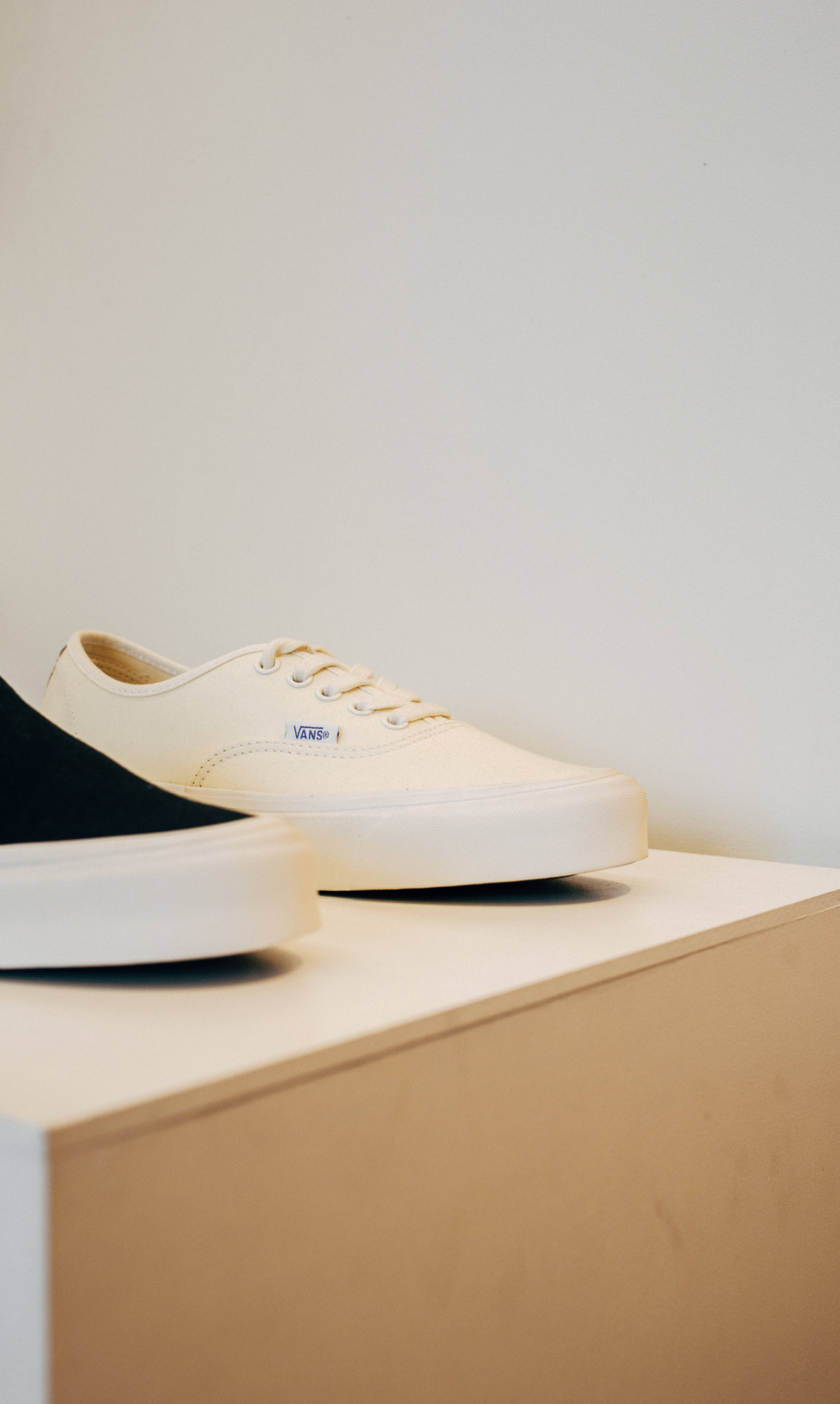 Vans 桌布 壁紙 — 兩雙極簡主義運動鞋，一雙奶油色一雙黑色，放在木塊上，背景是純白牆壁。