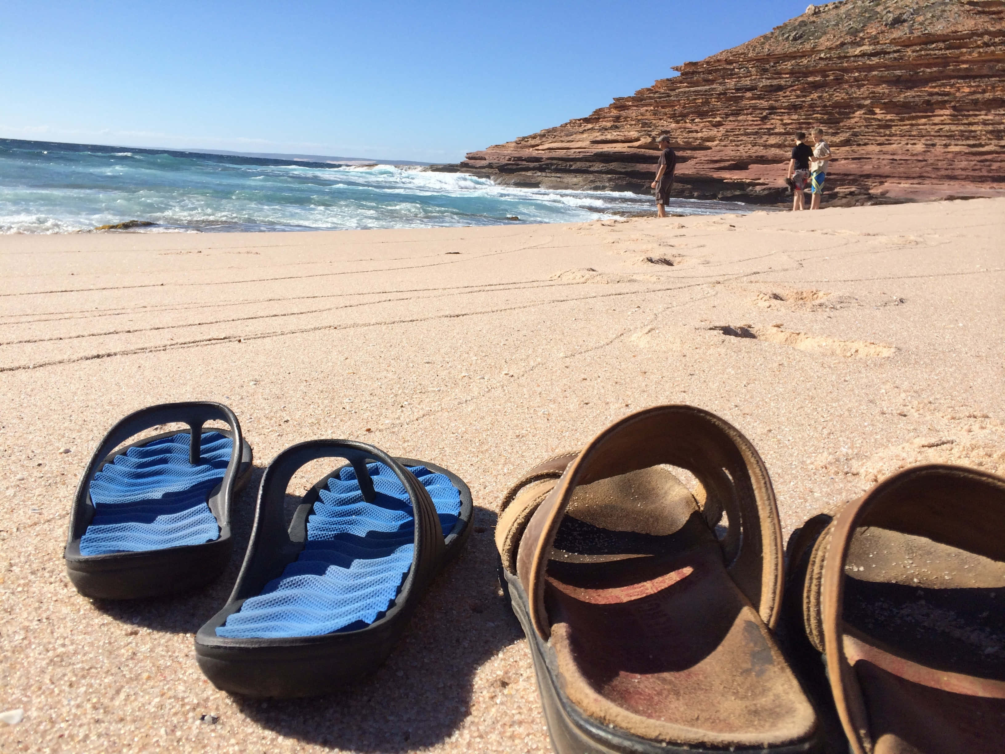 Beach Sandals 桌布 壁紙 — 兩雙涼鞋放在陽光普照的沙灘上，海浪拍打著紅色懸崖，遠處有人走動。