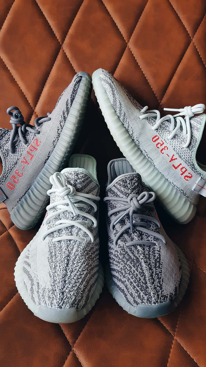 Yeezy 桌布 壁紙 — 兩雙灰色針織Yeezy運動鞋，帶有紅色文字，放置在棕色絎縫皮革表面上。